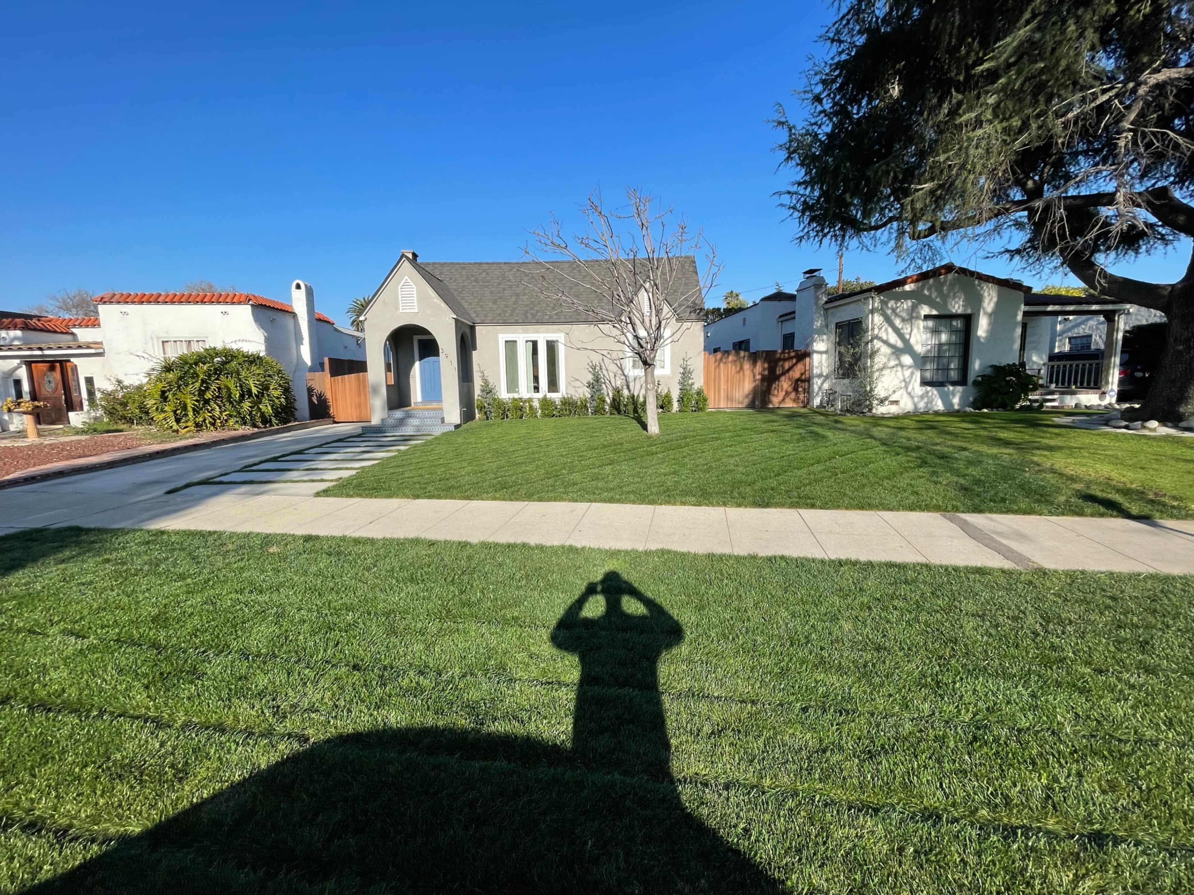 A house with a gray roof and white walls sits on a lawn in front of two neighboring buildings under a blue sky.