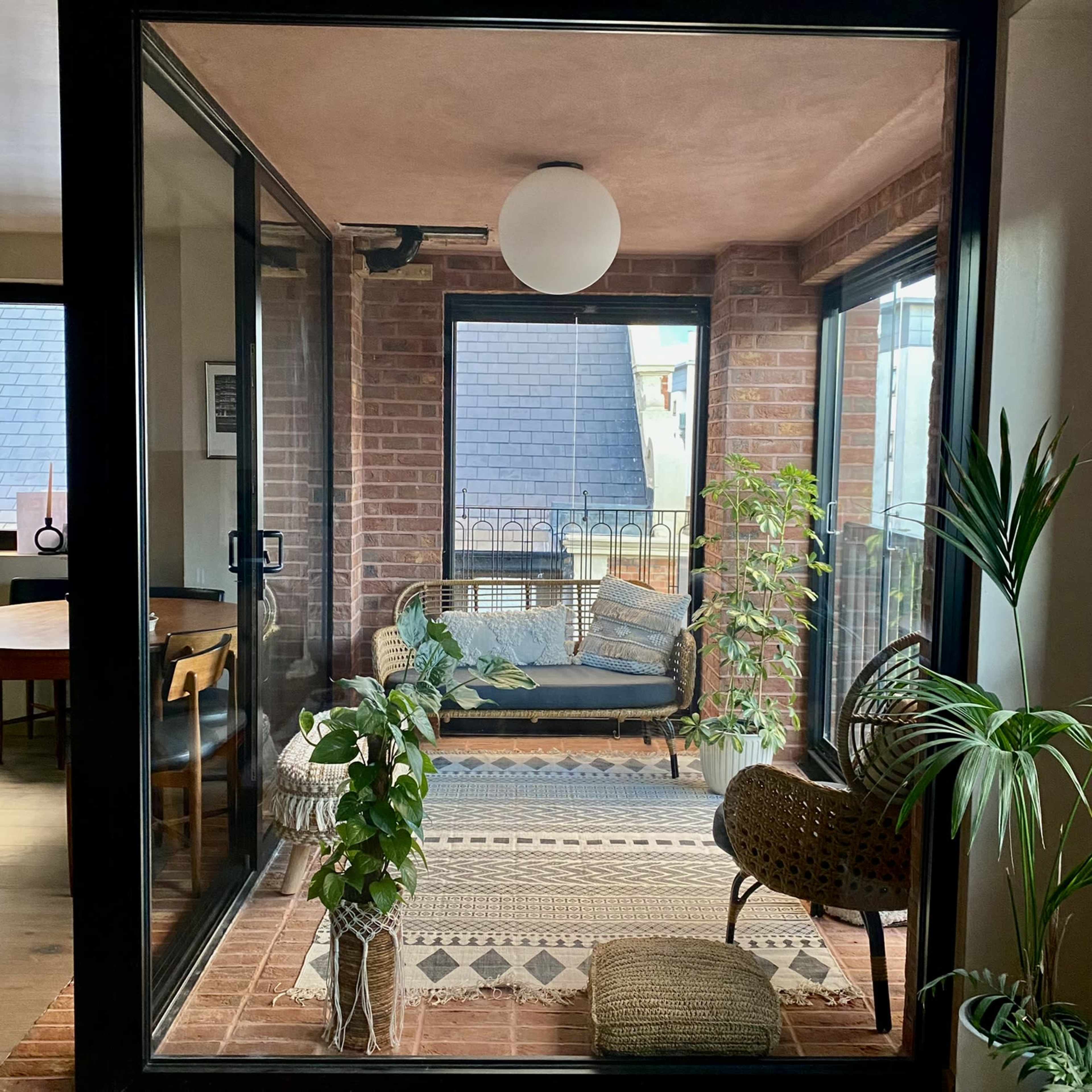 A cozy indoor seating area with large windows, featuring a rattan couch, plants, and a geometric-patterned rug.