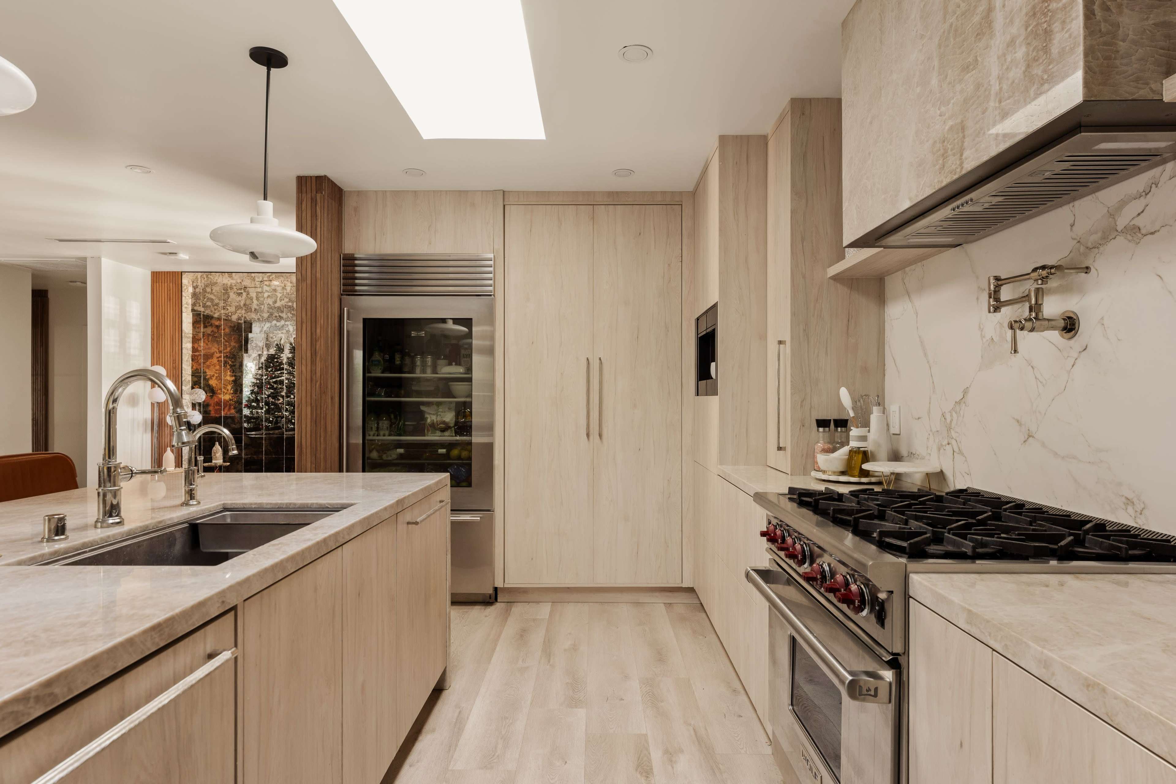 The image shows a modern kitchen featuring wood cabinetry, a large stainless steel refrigerator, and a gas range on a marble countertop.