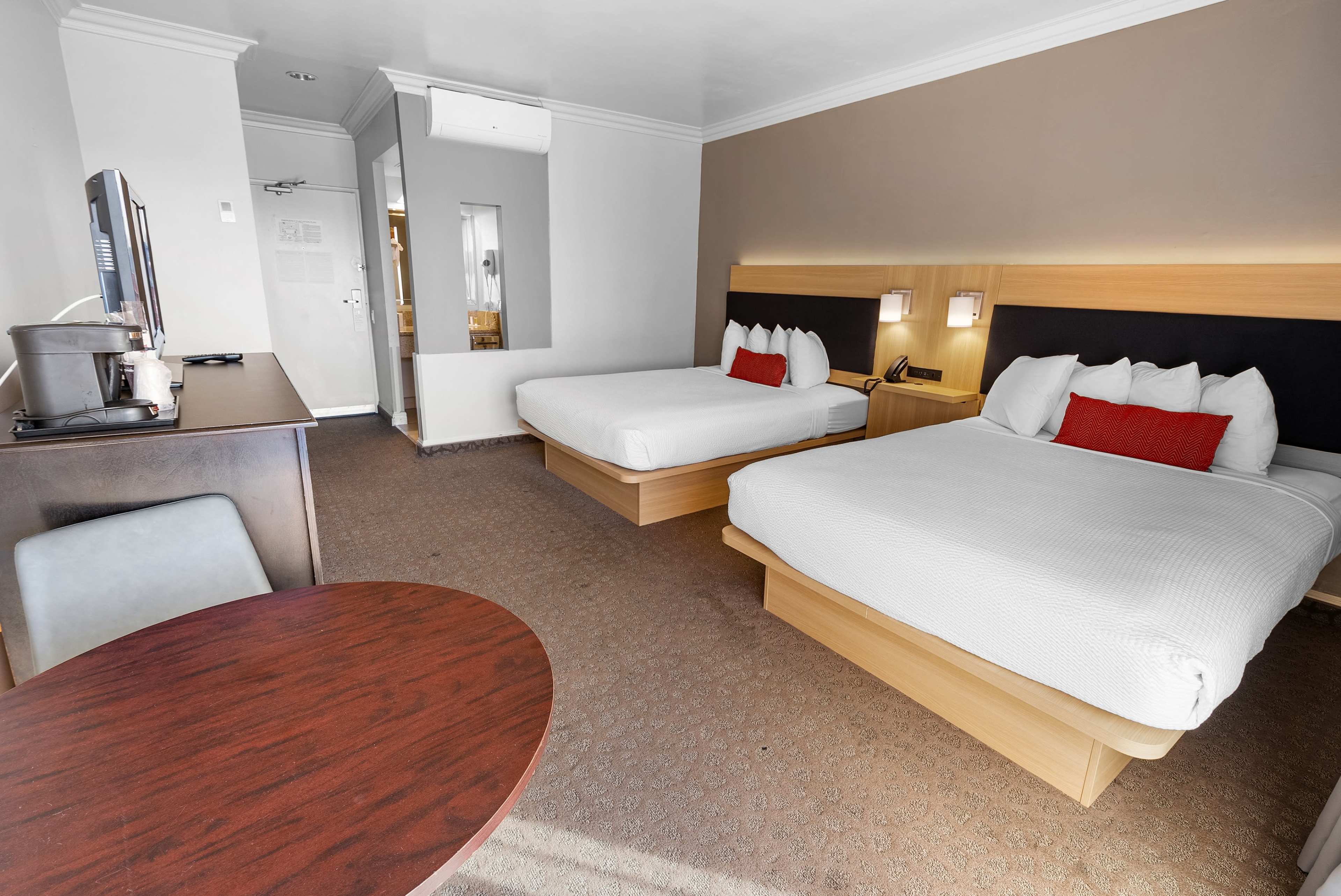 A hotel room with two double beds, a small round table, a TV unit, and a doorway leading to a bathroom.