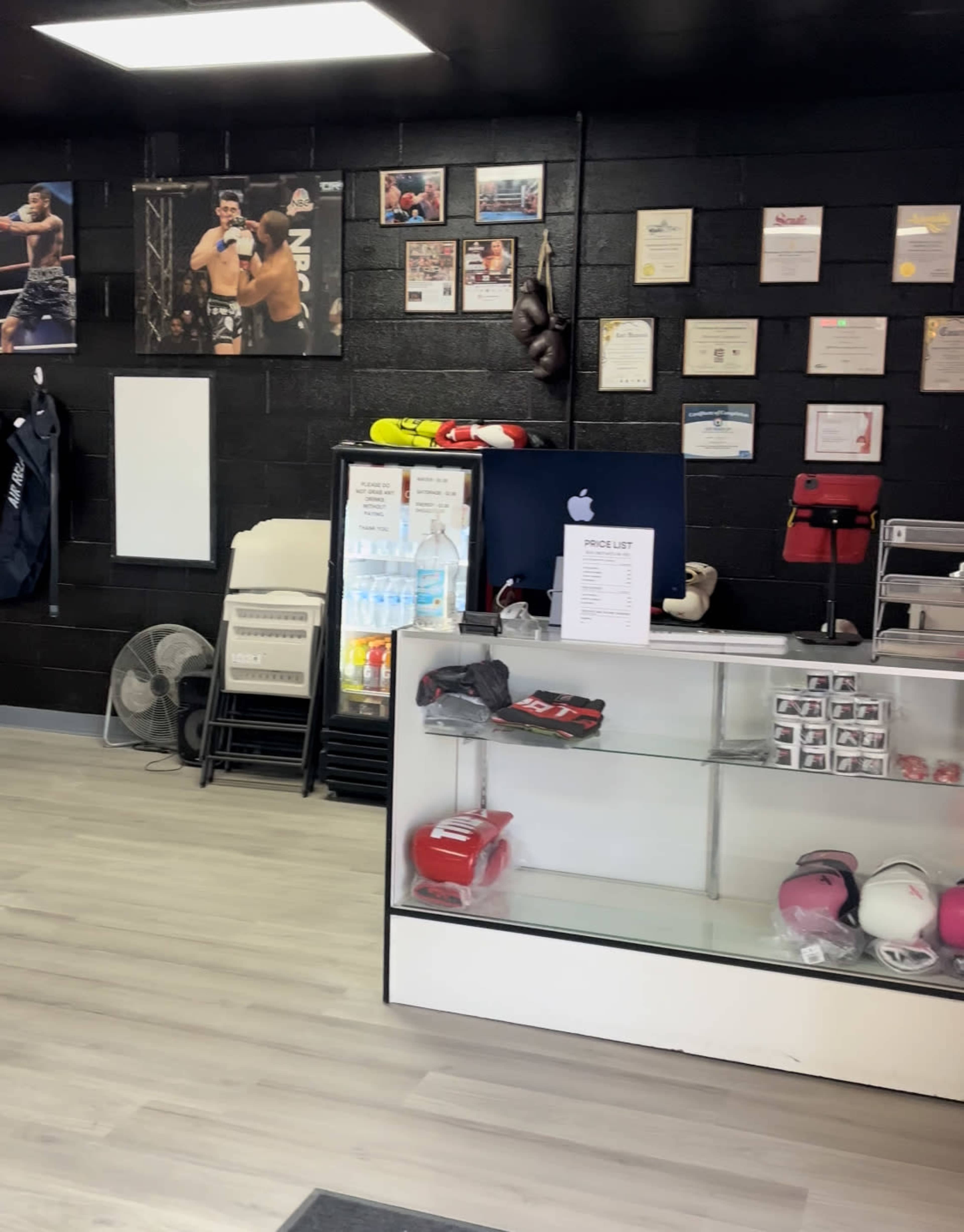 The image shows the interior of a boxing gym with a reception area featuring a display cabinet, a price list, and various boxing equipment on the counter.