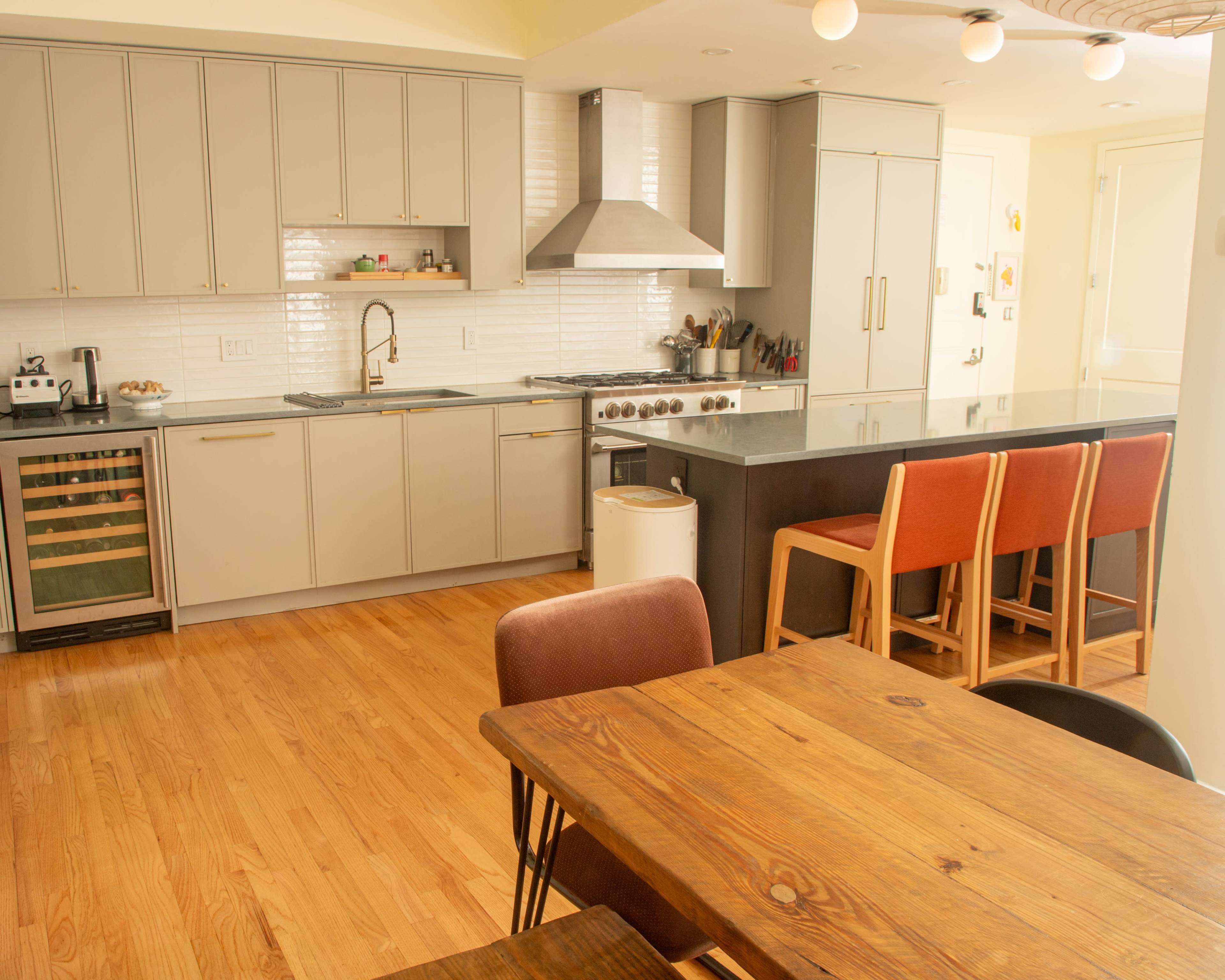 The image shows a modern kitchen with gray cabinets, a stainless steel stovetop, and a dining area featuring a wooden table and colorful chairs.