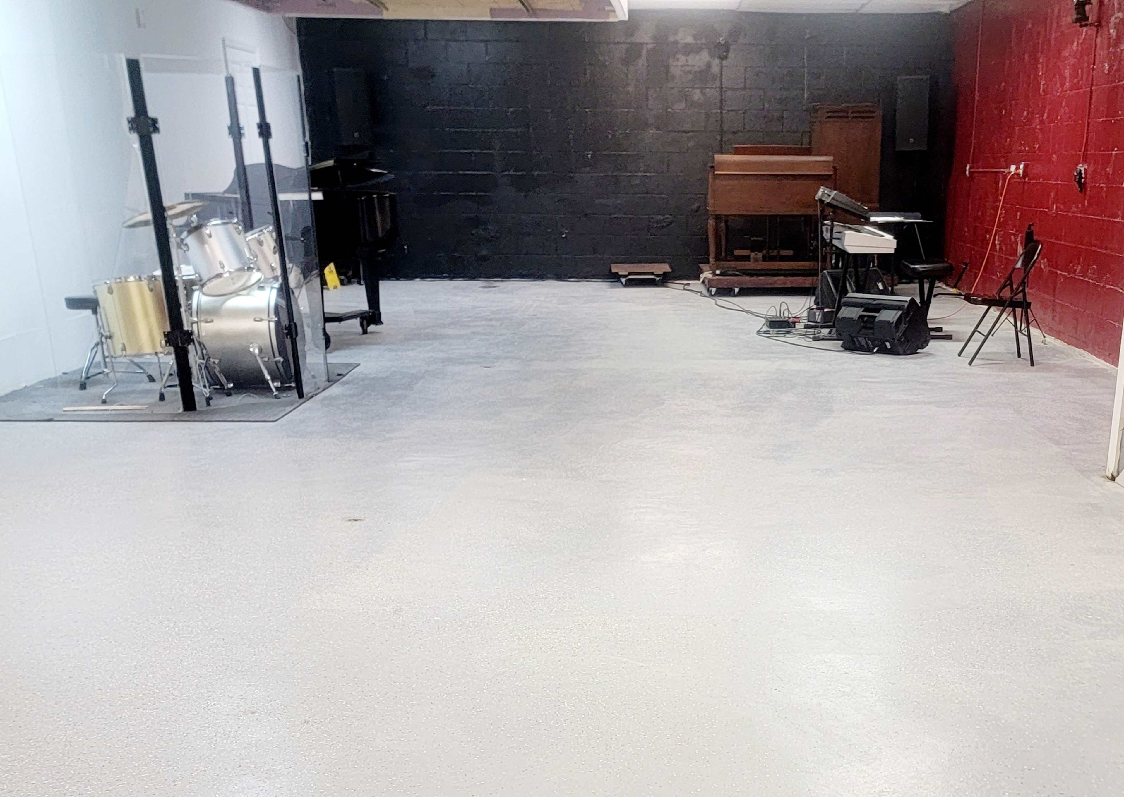 A large, empty music studio space with instruments including a drum set, piano, and electronic keyboard arranged against red and black walls.