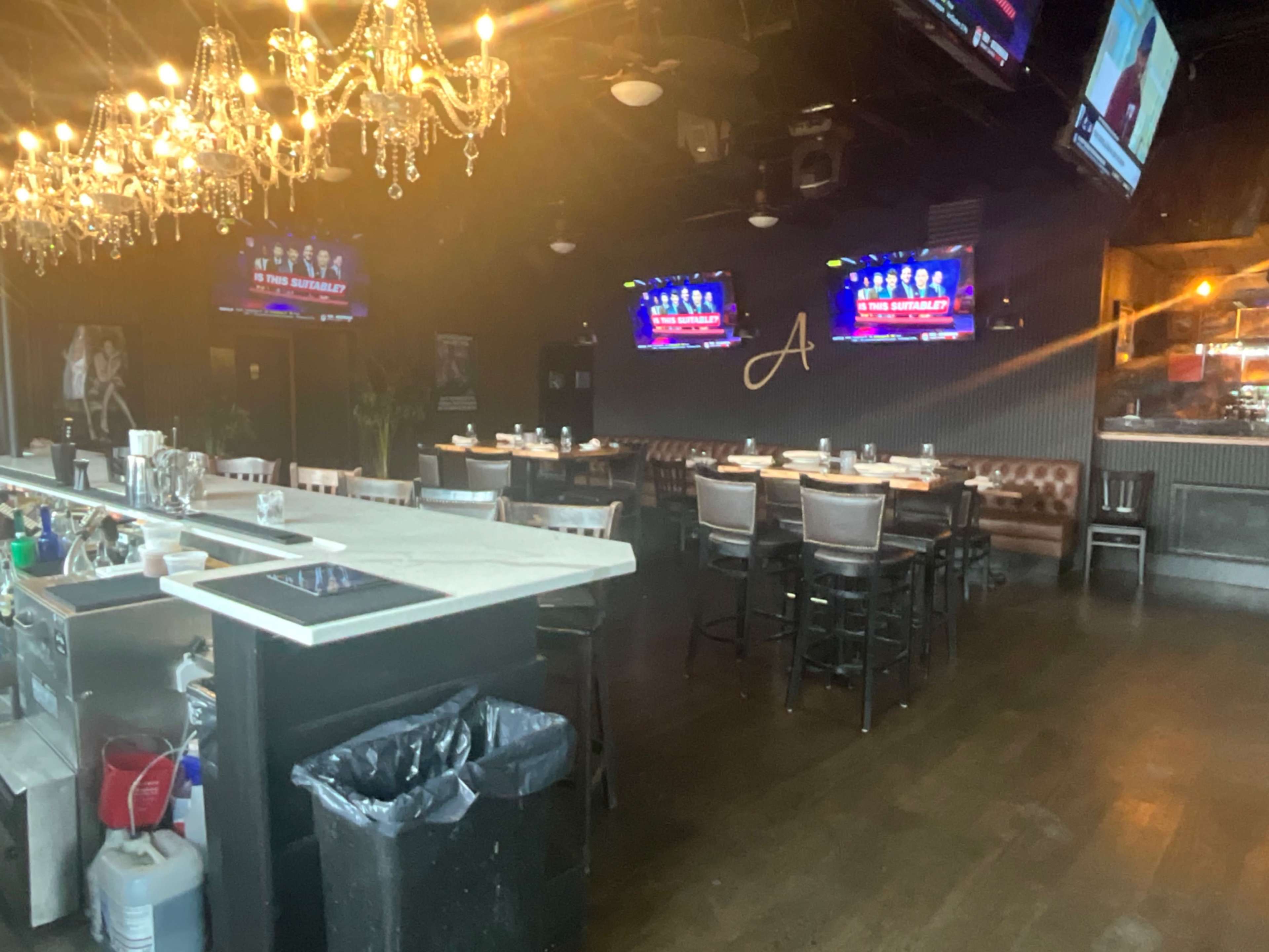 The image shows the interior of a restaurant featuring a bar area with a chandelier, seating arrangements, and multiple mounted televisions displaying content.