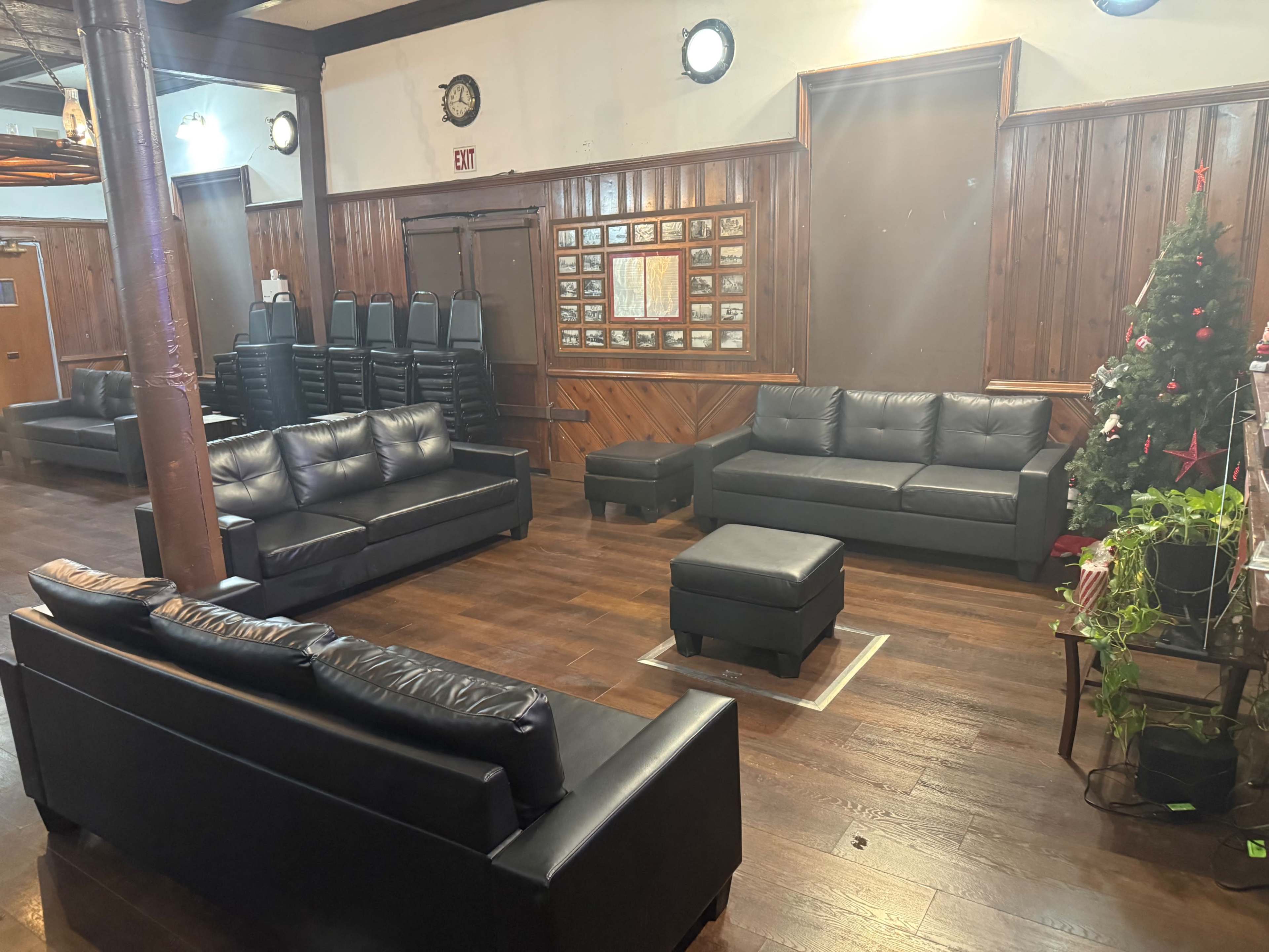 The image shows a spacious room with several black leather sofas, a coffee table, and a Christmas tree in one corner, along with stacked chairs against the wall.