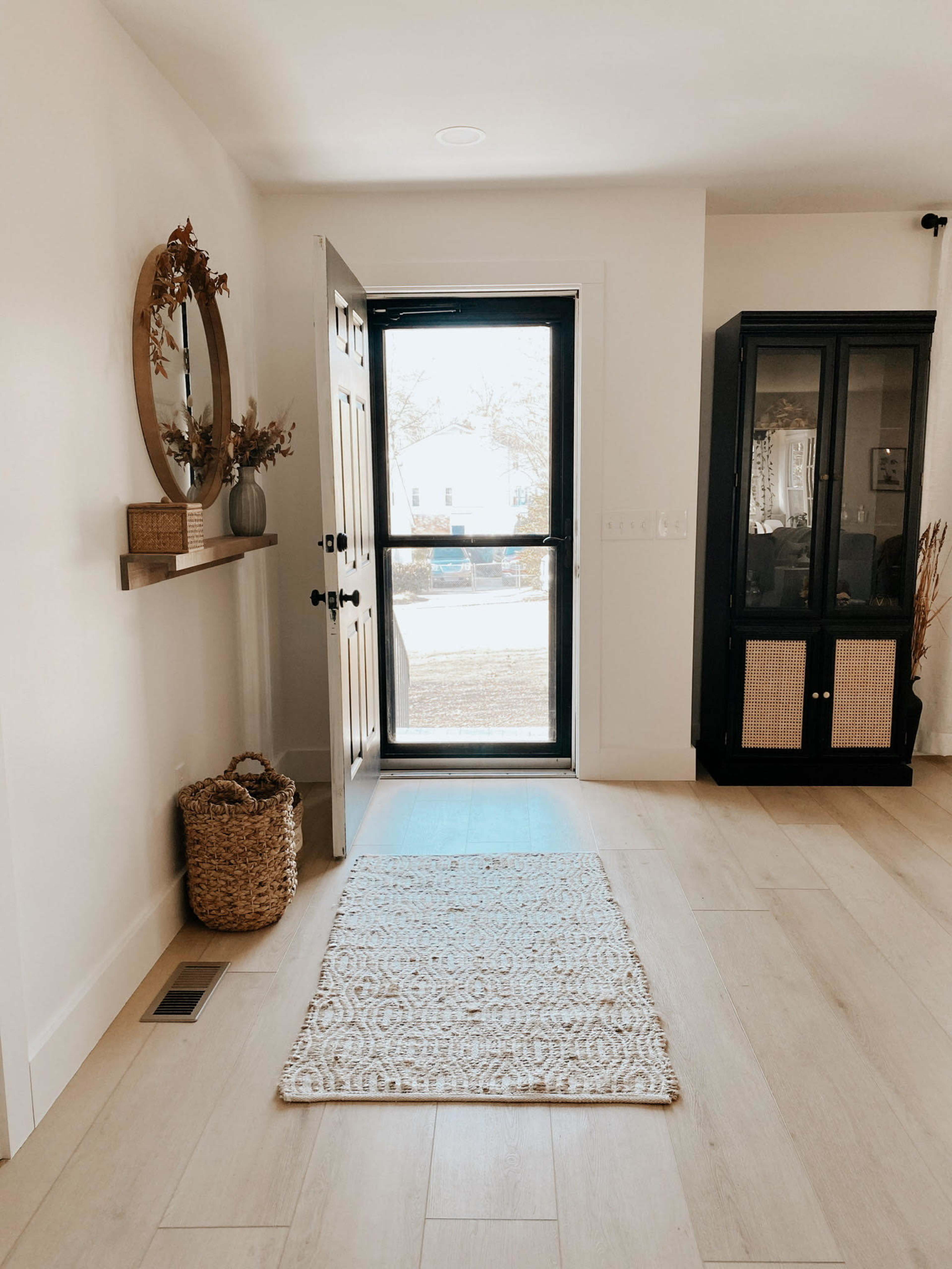 A brightly lit entryway with a woven rug, a round mirror, a decorative shelf, and a glass-paned door leading outside.