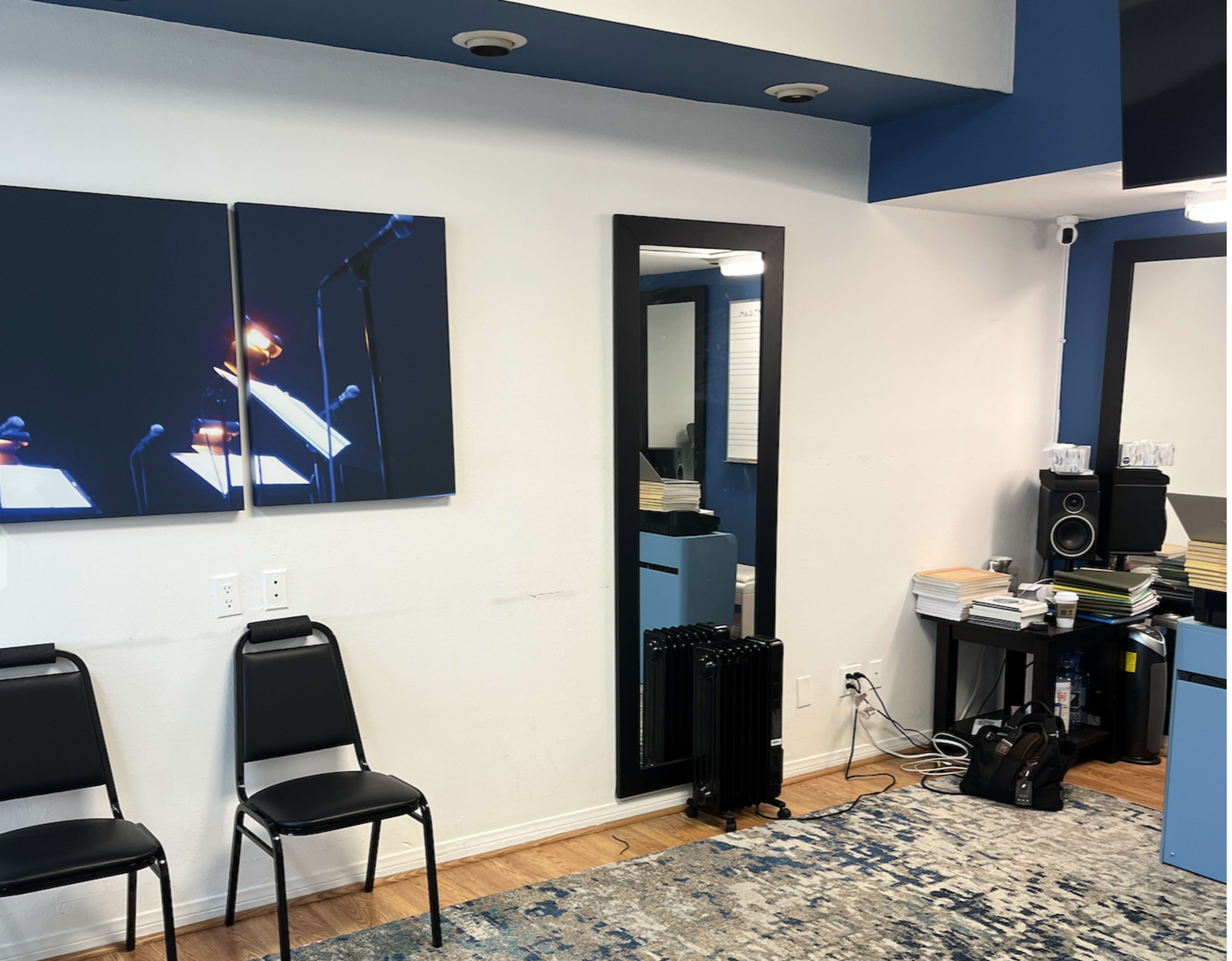 A room with two black chairs, a wall-mounted mirror, an abstract artwork, and a cluttered area with books and equipment.
