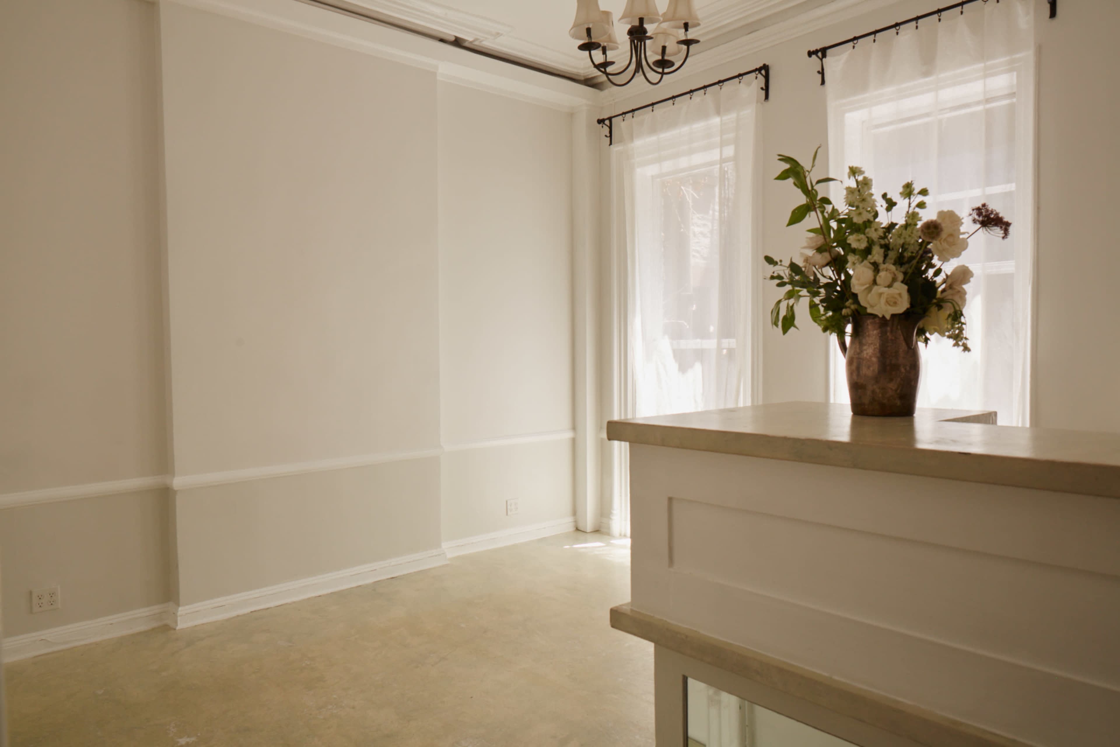 A light-filled room with pale walls, large windows covered by sheer curtains, and a decorative vase of flowers on a countertop.
