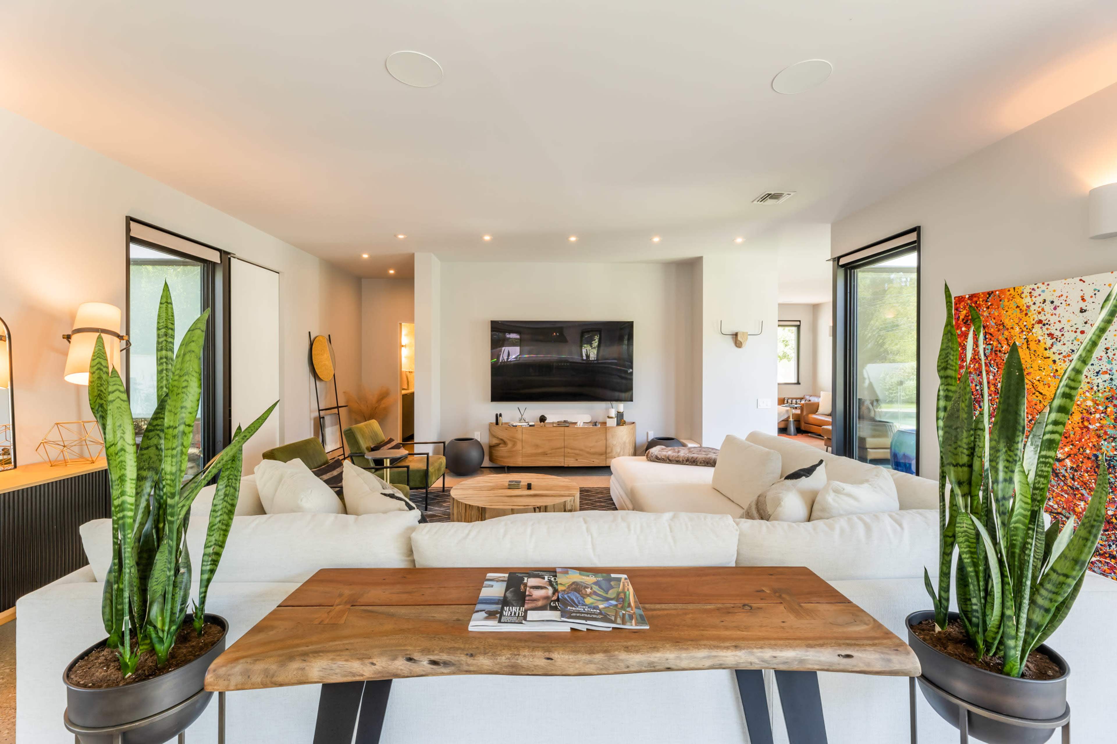 A modern living room featuring a white sofa, a wooden coffee table, indoor plants, and a large wall-mounted television.