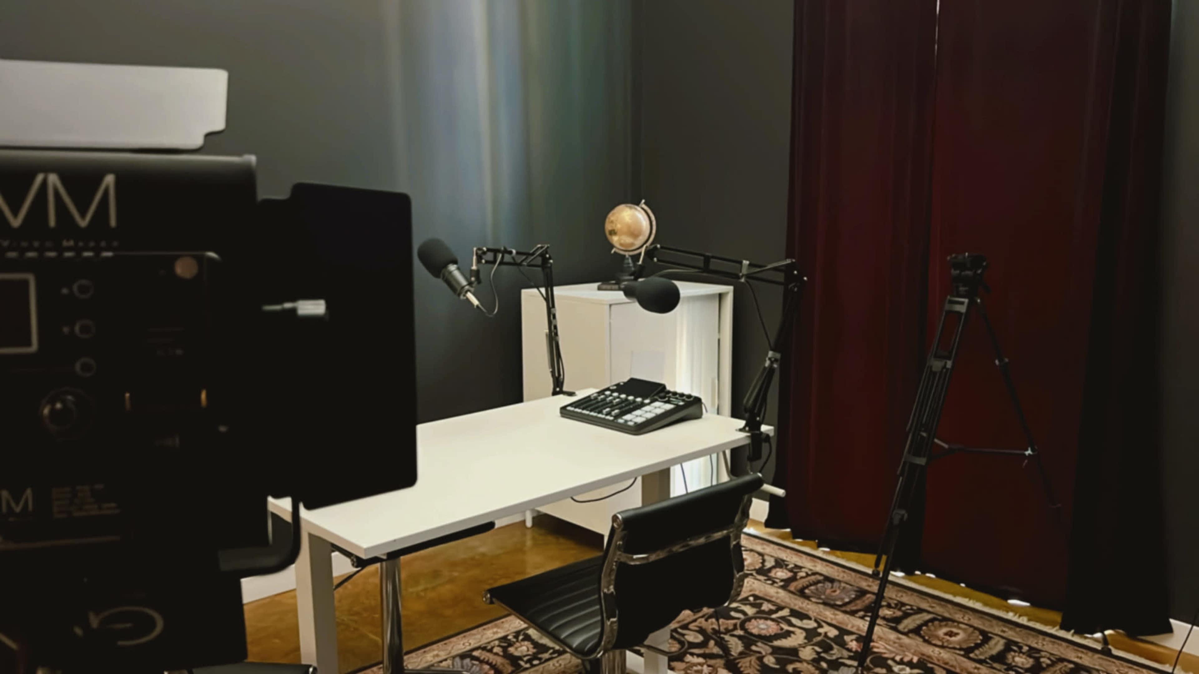A minimalistic recording studio setup with a white desk, a microphone on an arm, a camera tripod, and a globe lamp against a dark backdrop.
