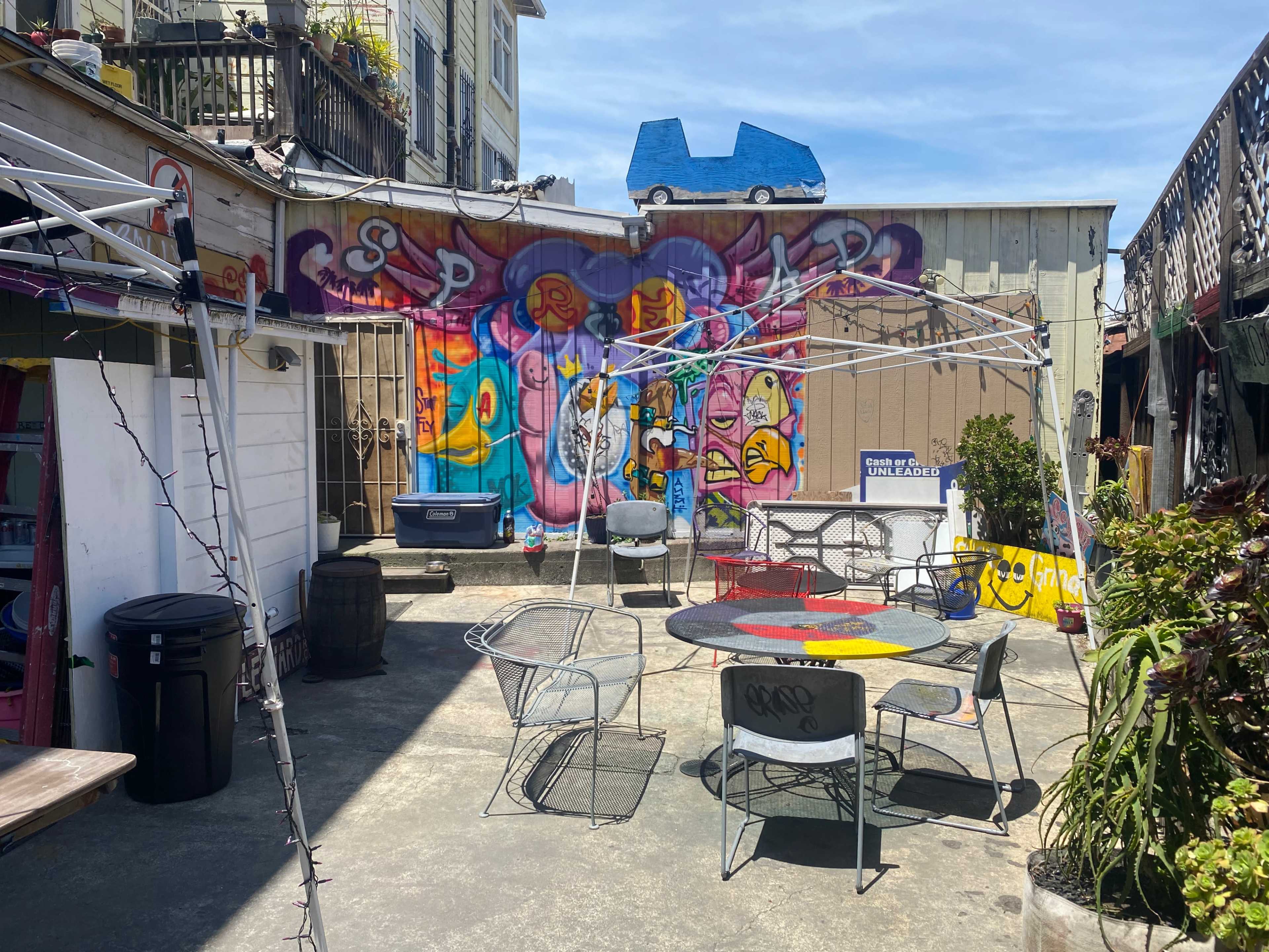 The scene shows a courtyard with metal chairs and tables, surrounded by colorful graffiti murals on the walls and a partially dismantled white canopy overhead.