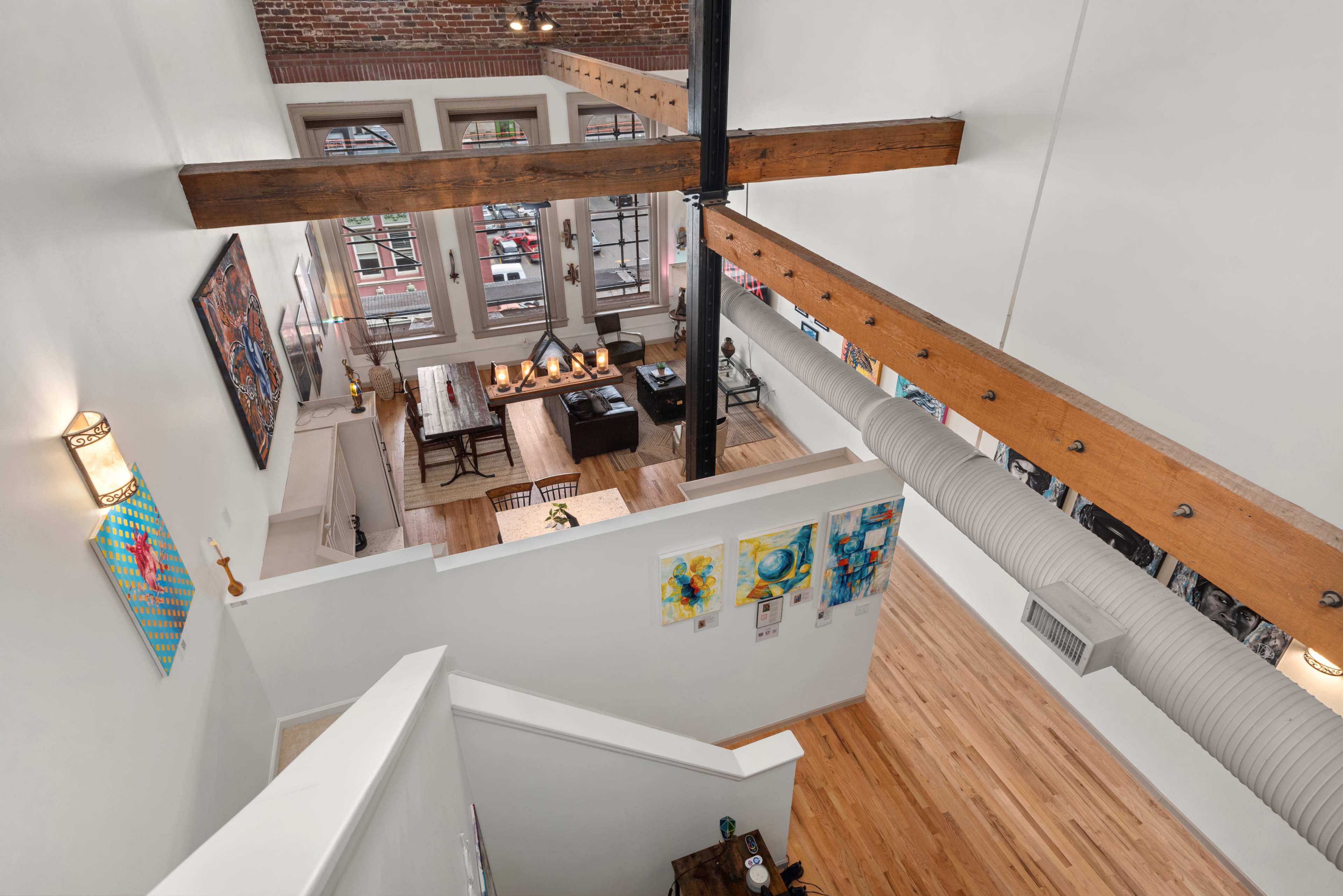 The image shows the interior of a loft-style apartment with exposed brick walls, wooden beams, and a high ceiling, featuring an open living area and dining space.