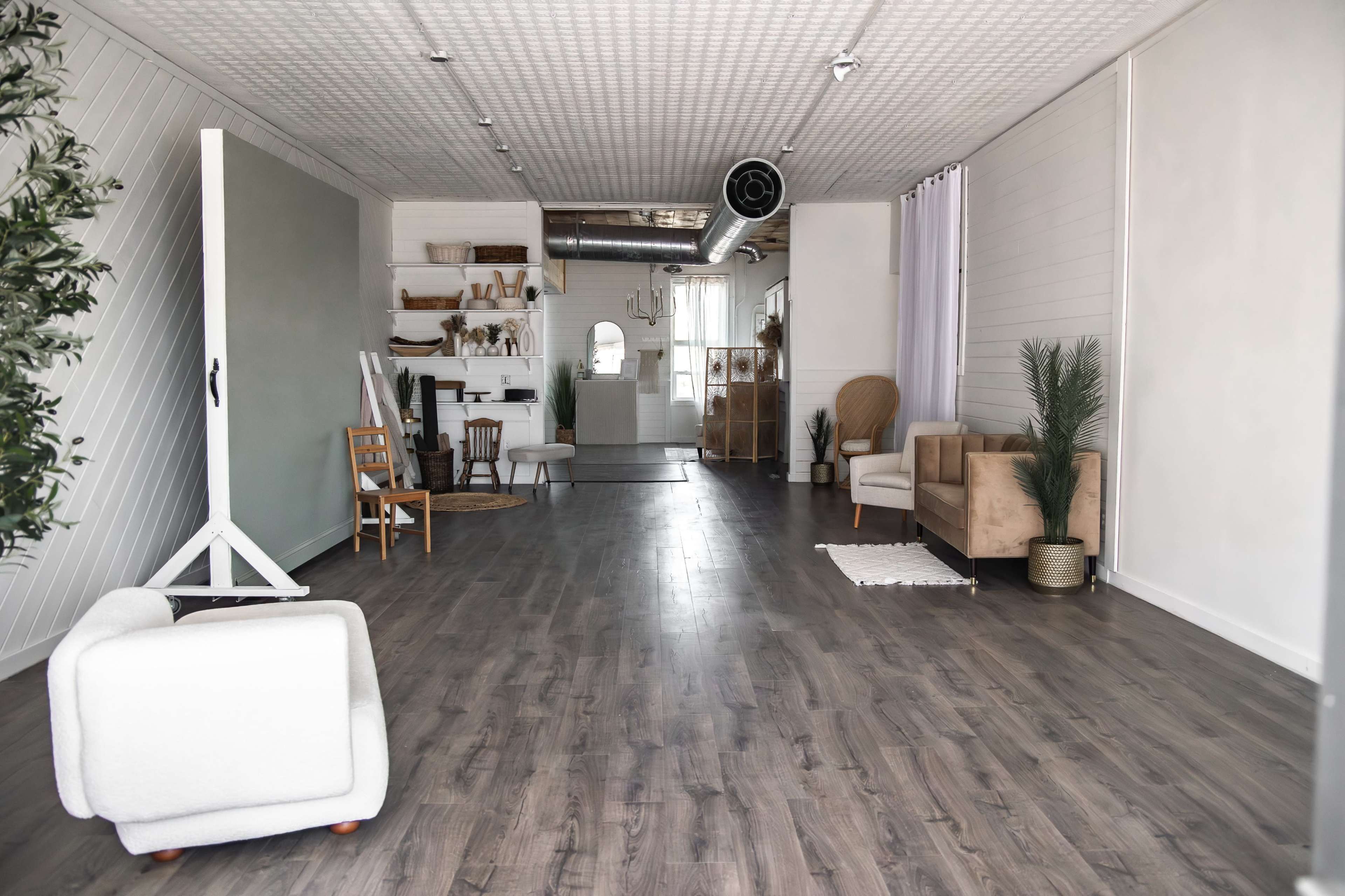 The image shows a spacious, modern studio with wooden flooring, minimalist furniture, and a well-lit area featuring a white backdrop and decorative plants.