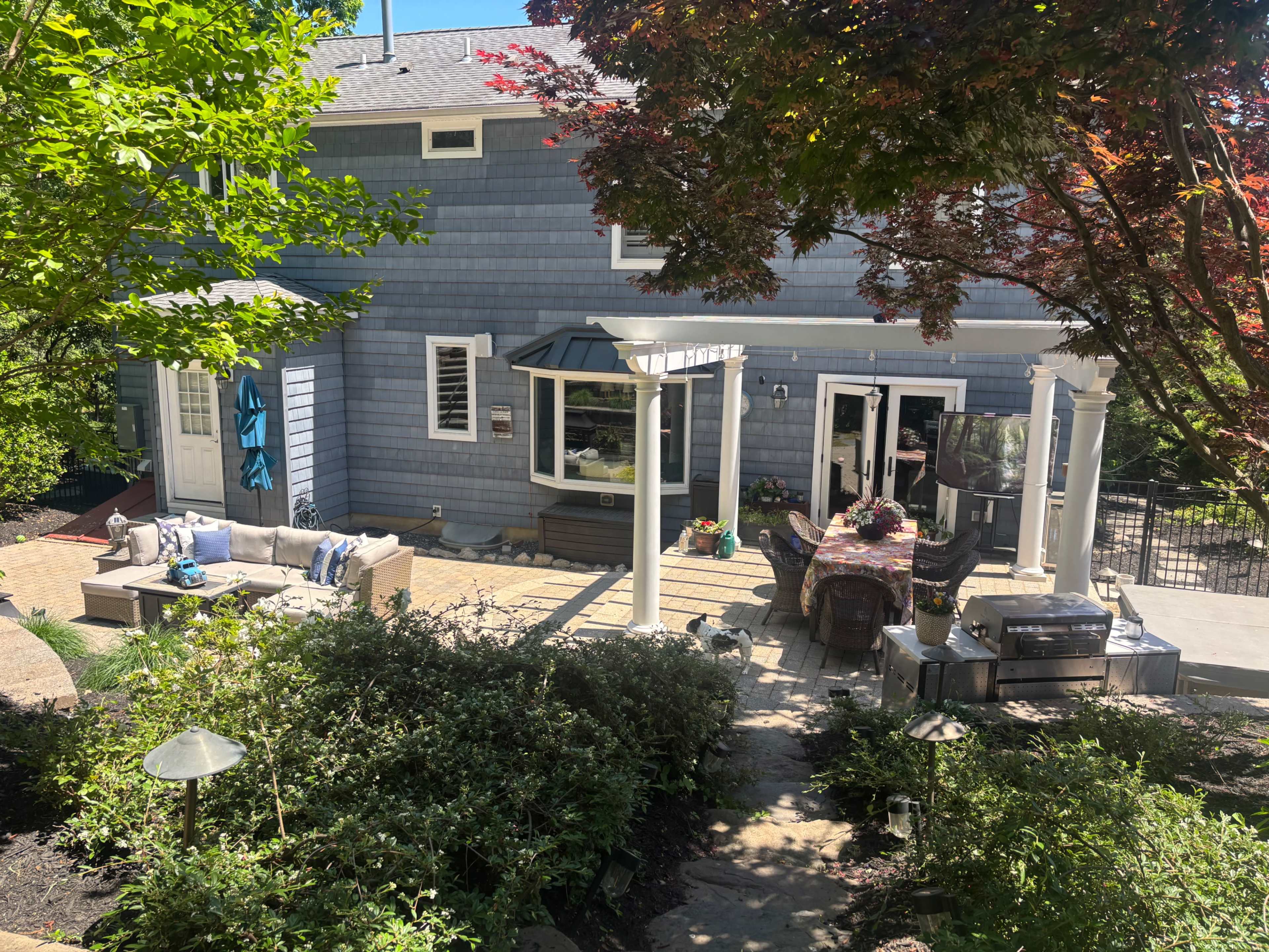 The image shows a backyard patio area with seating and a dining table, surrounded by greenery and featuring a blue house in the background.