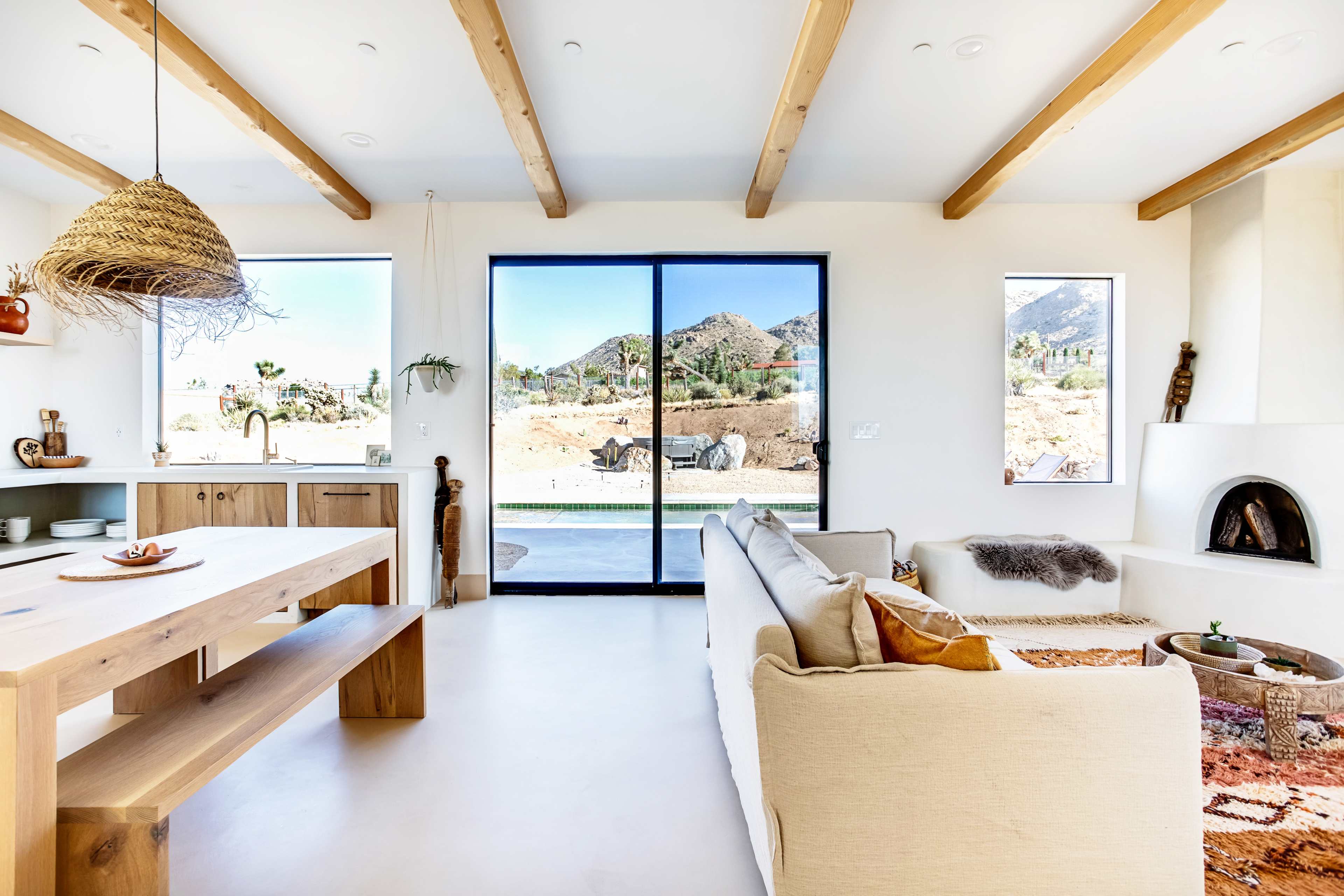 A modern living space features wooden beams, large windows overlooking a desert landscape, and a minimalist kitchen and seating area.