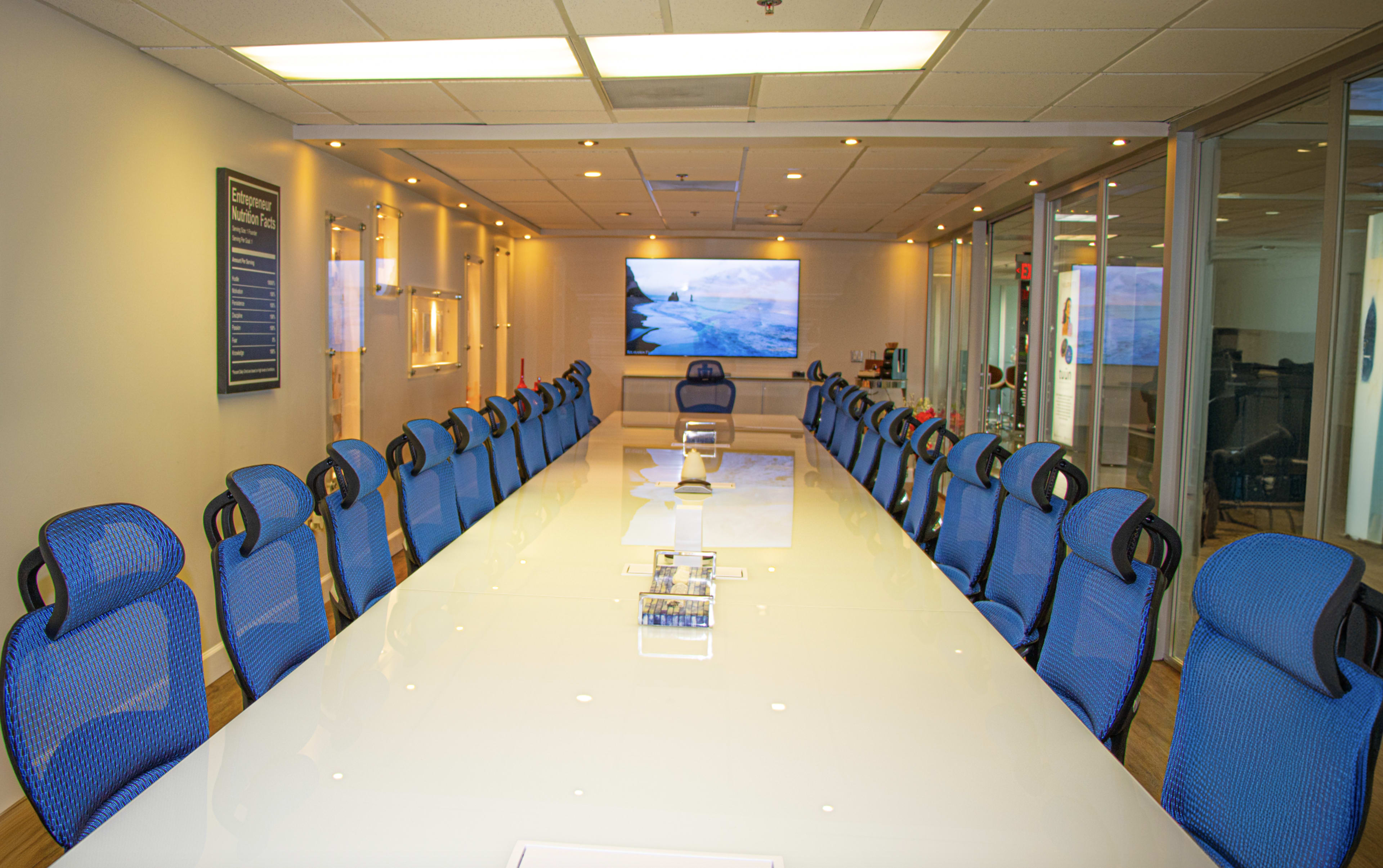 A large, modern conference room features a long white table surrounded by blue ergonomic chairs and a screen displaying a landscape image on one wall.