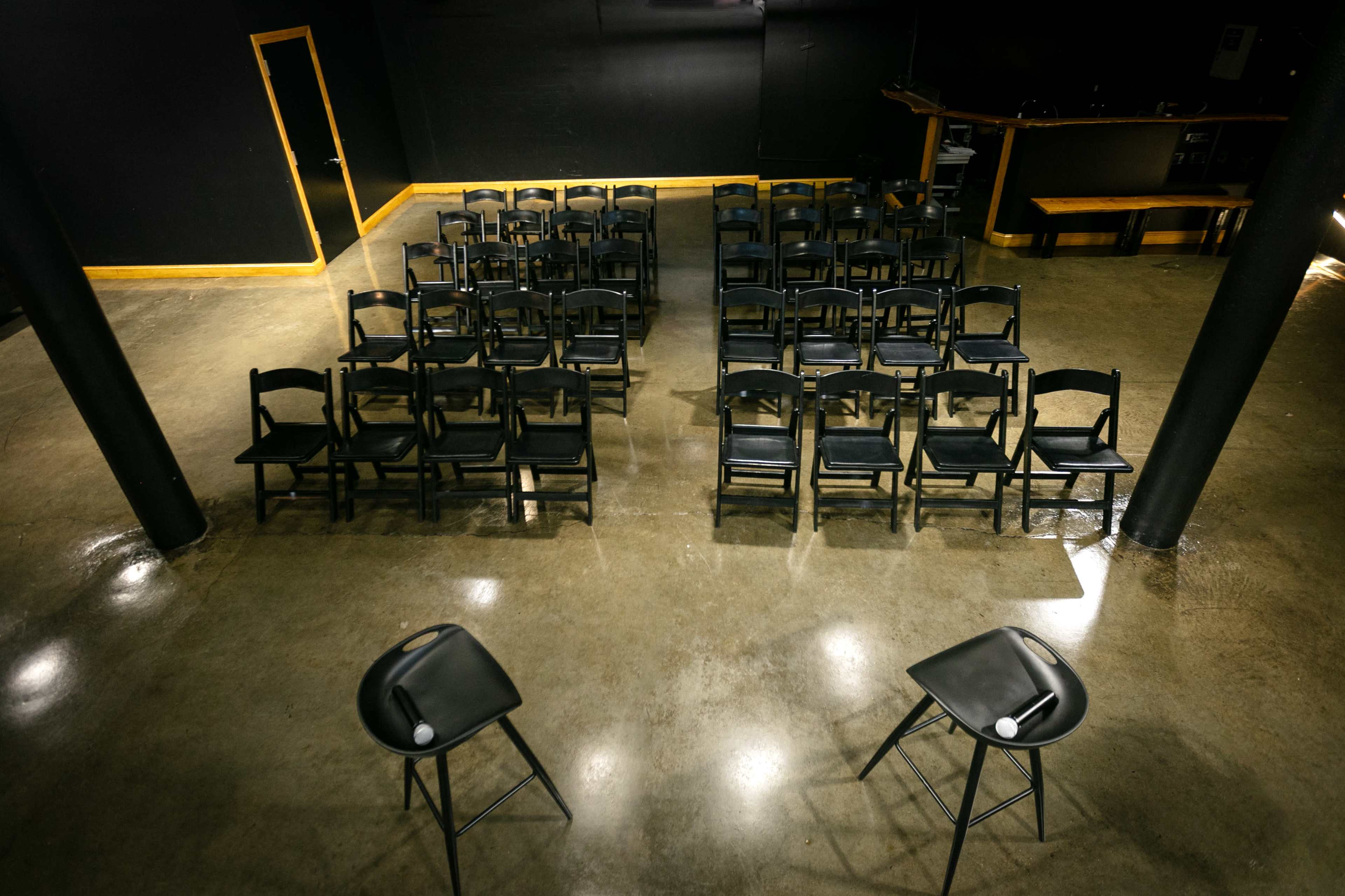 A row of black chairs is arranged in a sparse, empty venue with two high stools positioned in the foreground.
