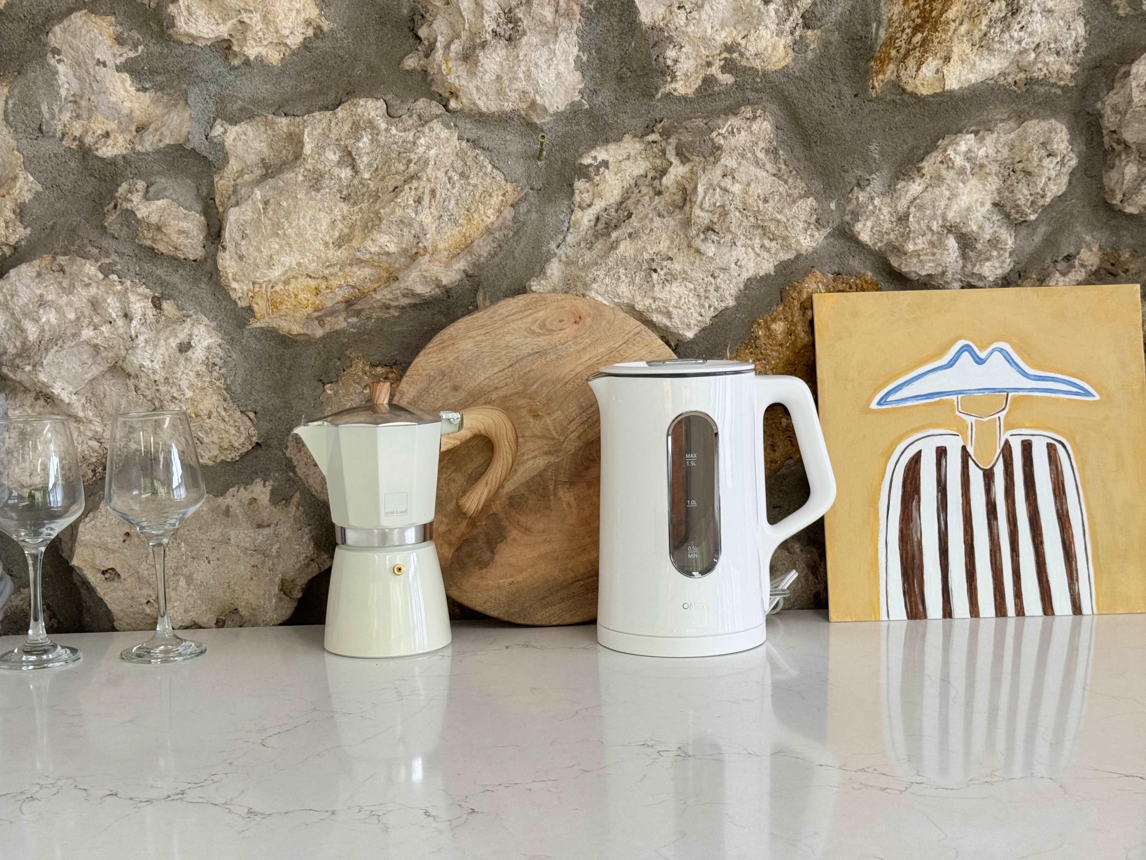 The image shows a countertop with a moka pot, an electric kettle, a wooden cutting board, two wine glasses, and a painting of a person in a hat against a stone wall.