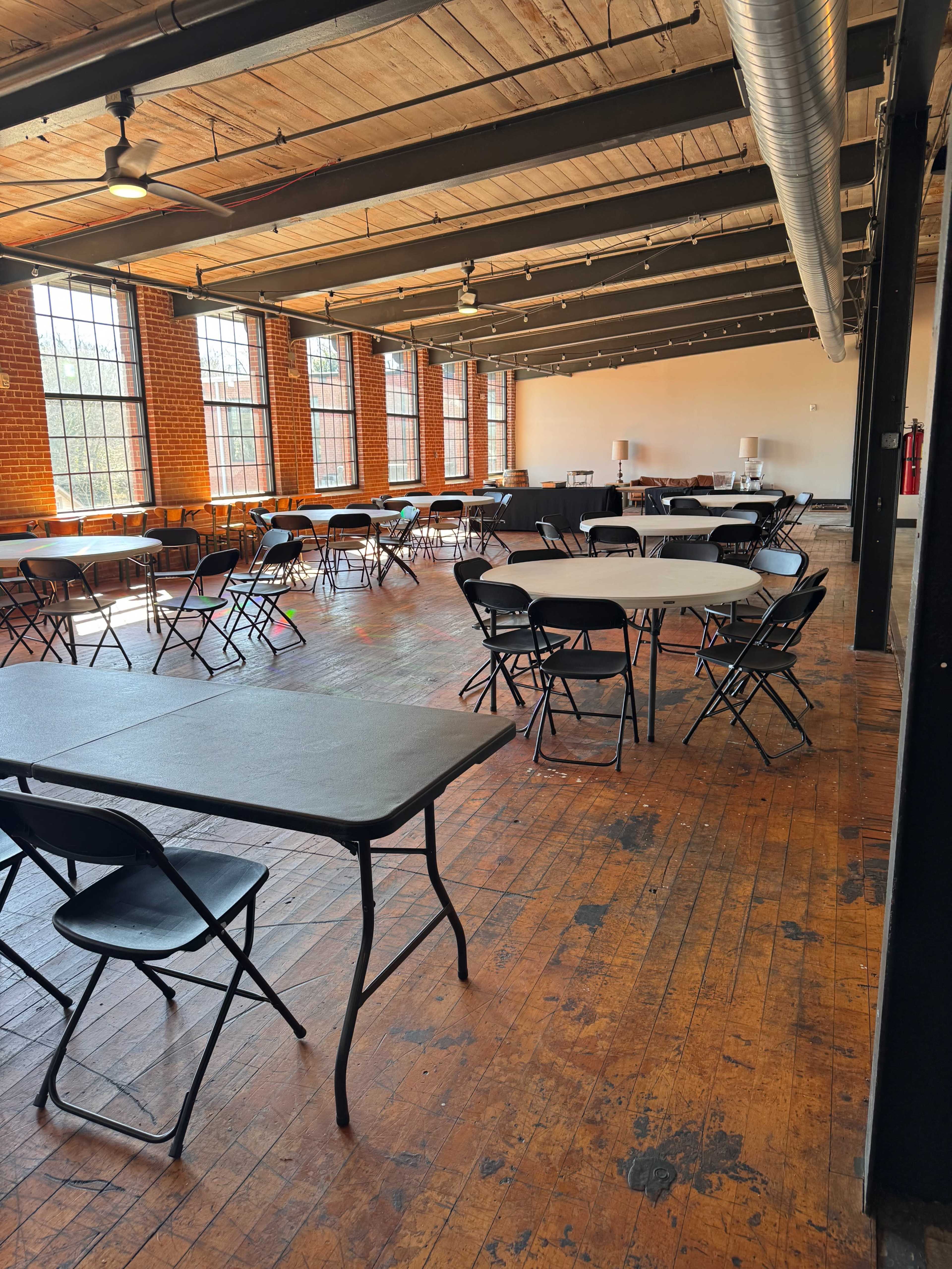 The image shows a spacious room with multiple round and rectangular tables set up, surrounded by black folding chairs, and illuminated by large windows.