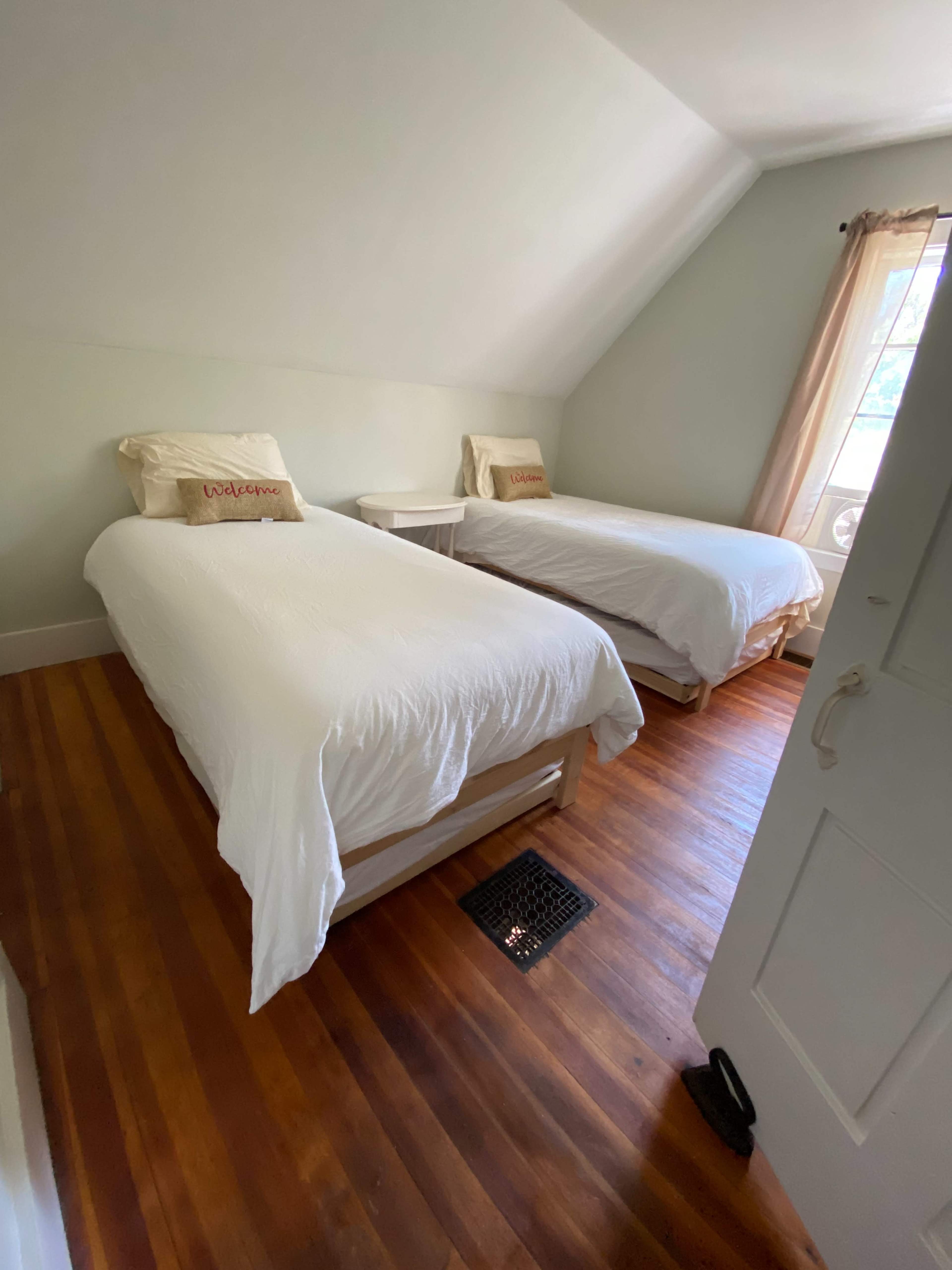A light-colored bedroom with two single beds, a nightstand, and a window allowing natural light.