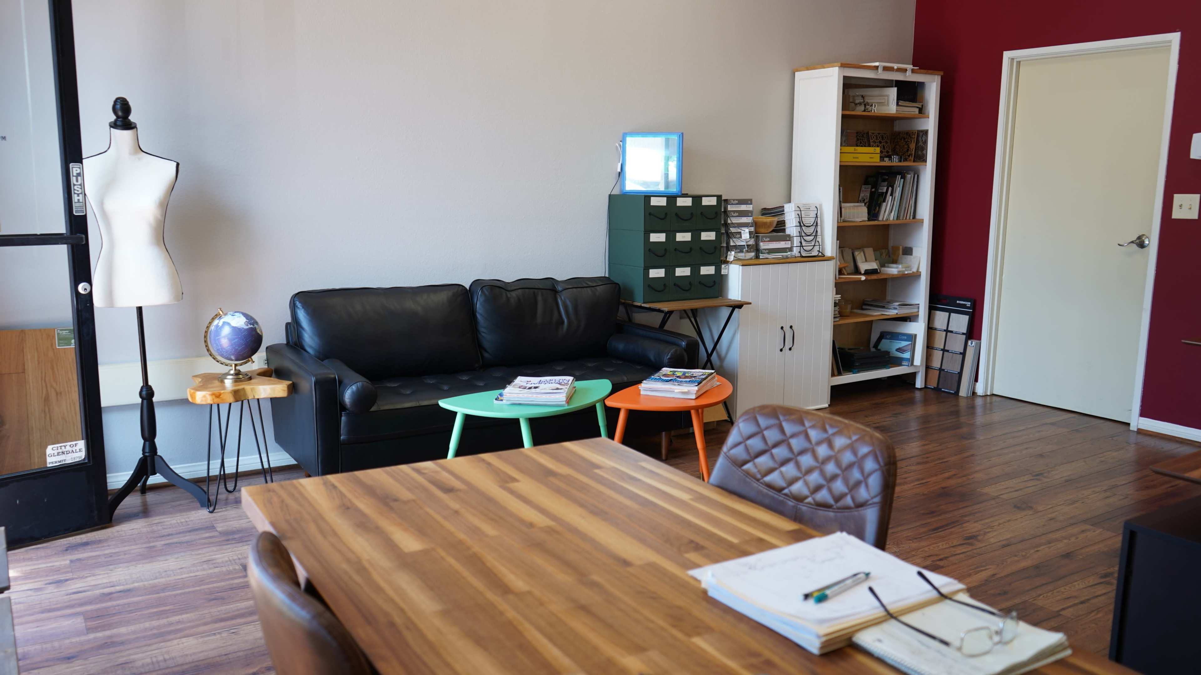The image depicts a modern interior space featuring a black leather couch, a wooden dining table, colorful side tables, and bookshelves filled with various items.