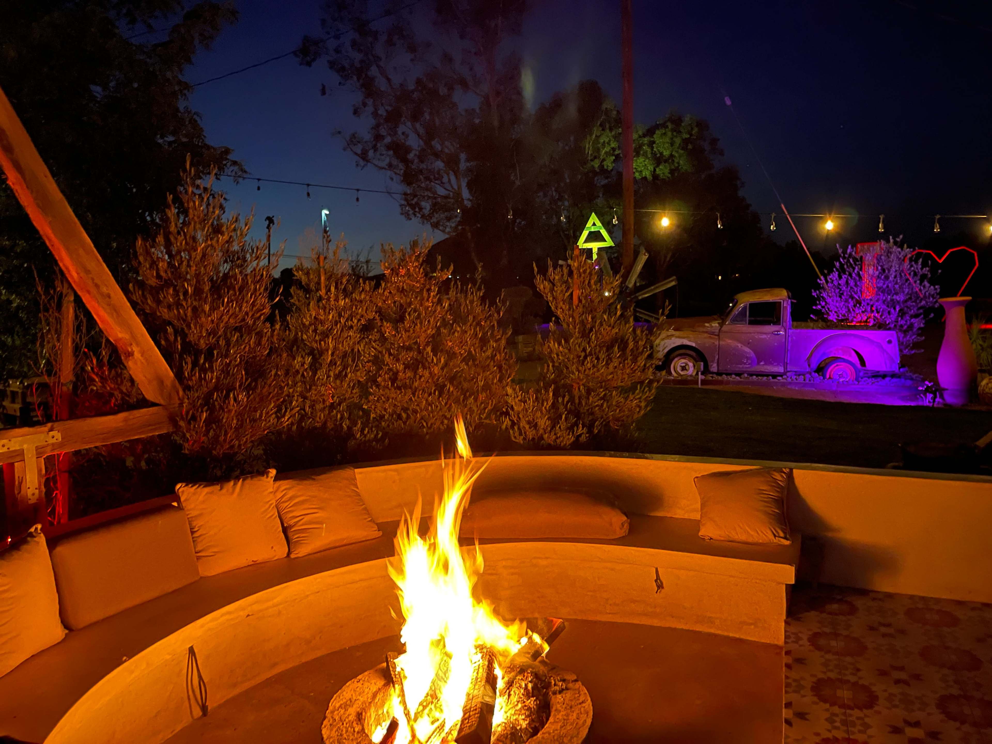 A colorful fire pit surrounded by seating illuminates a backyard at dusk, with an old truck and decorative lights visible in the background.