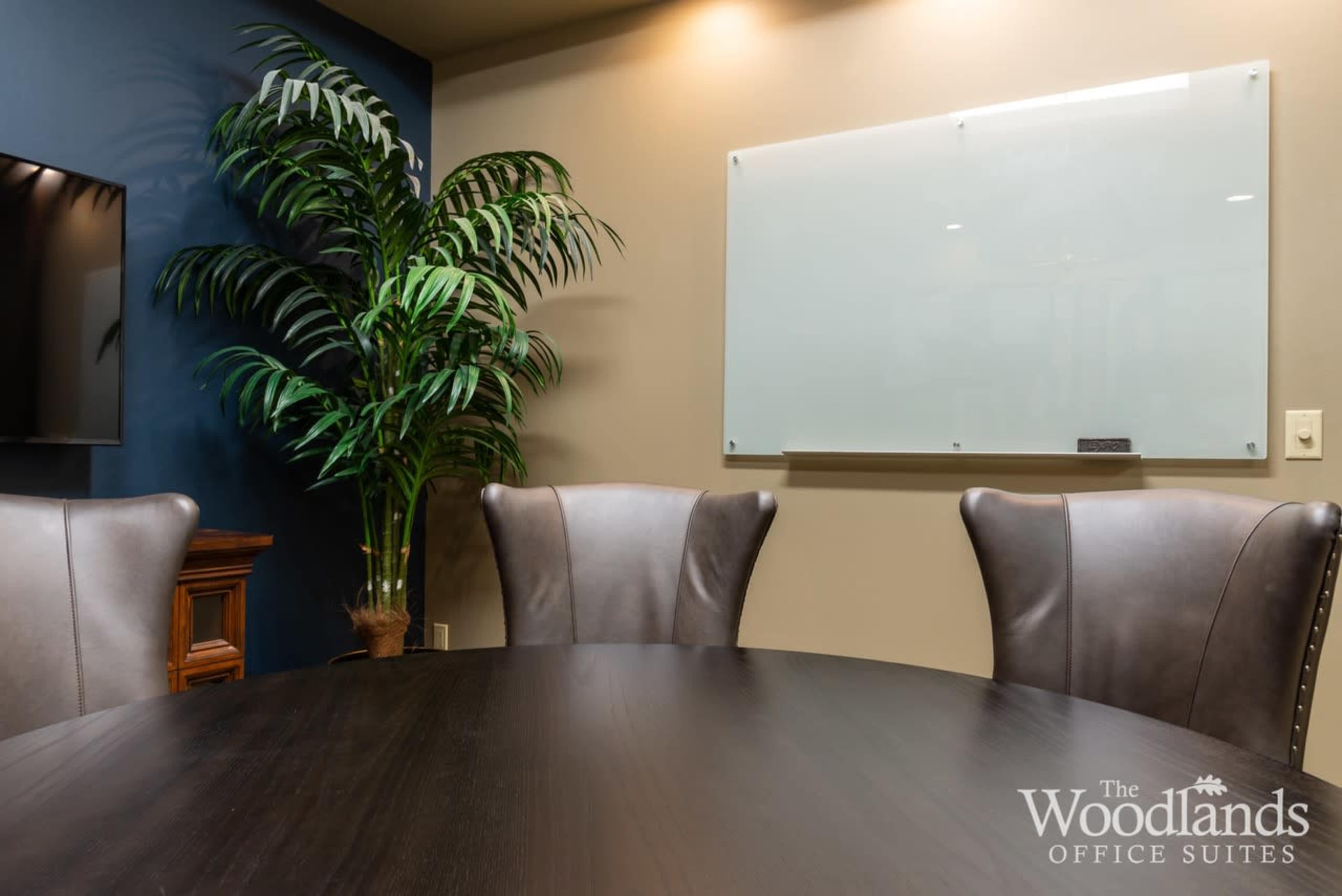 The image shows a conference room with a round wooden table, leather chairs, a potted palm plant, and a wall-mounted whiteboard.