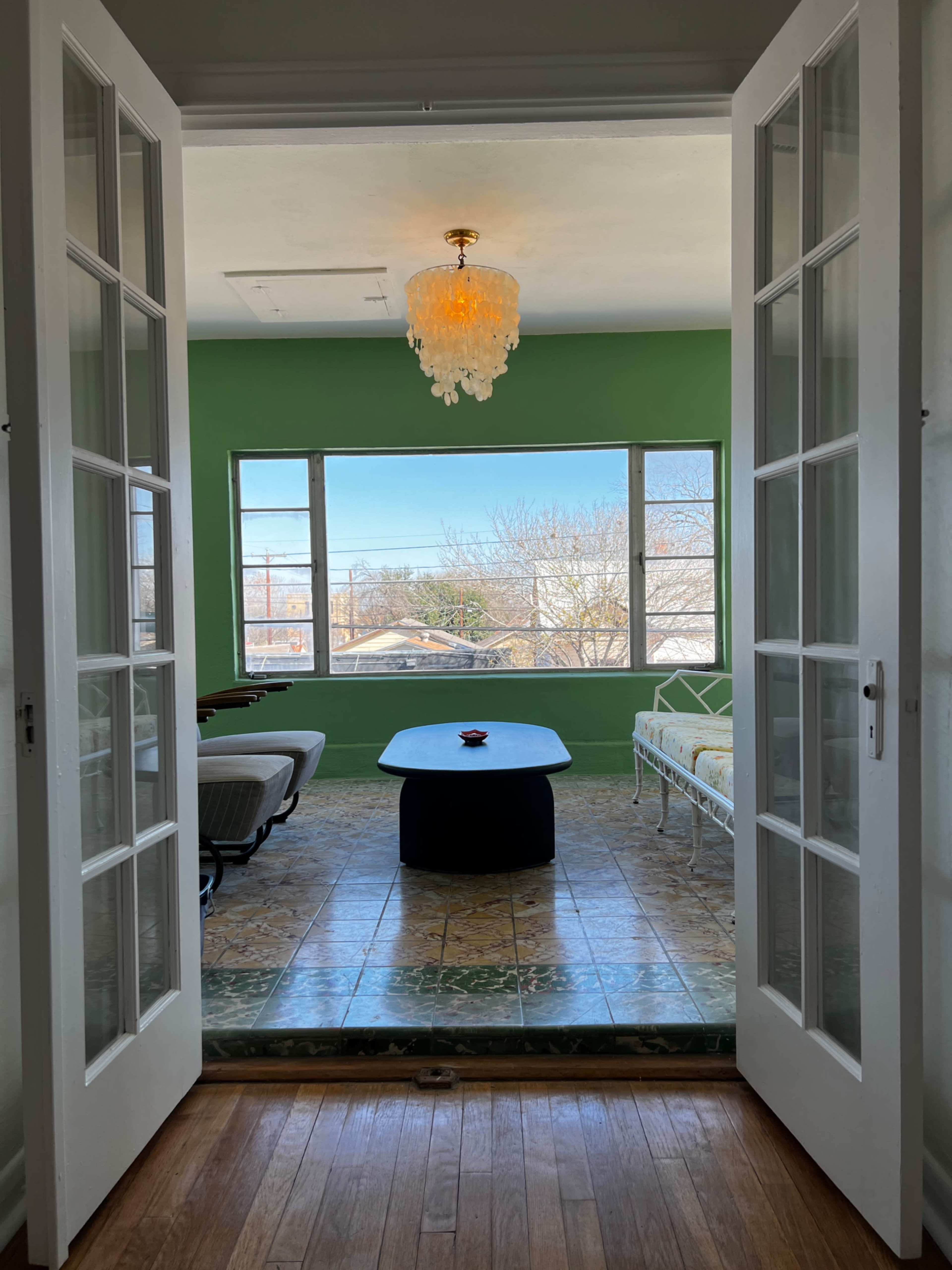 A pair of white French doors opens to a bright room with a green wall, large windows, and a chandelier.