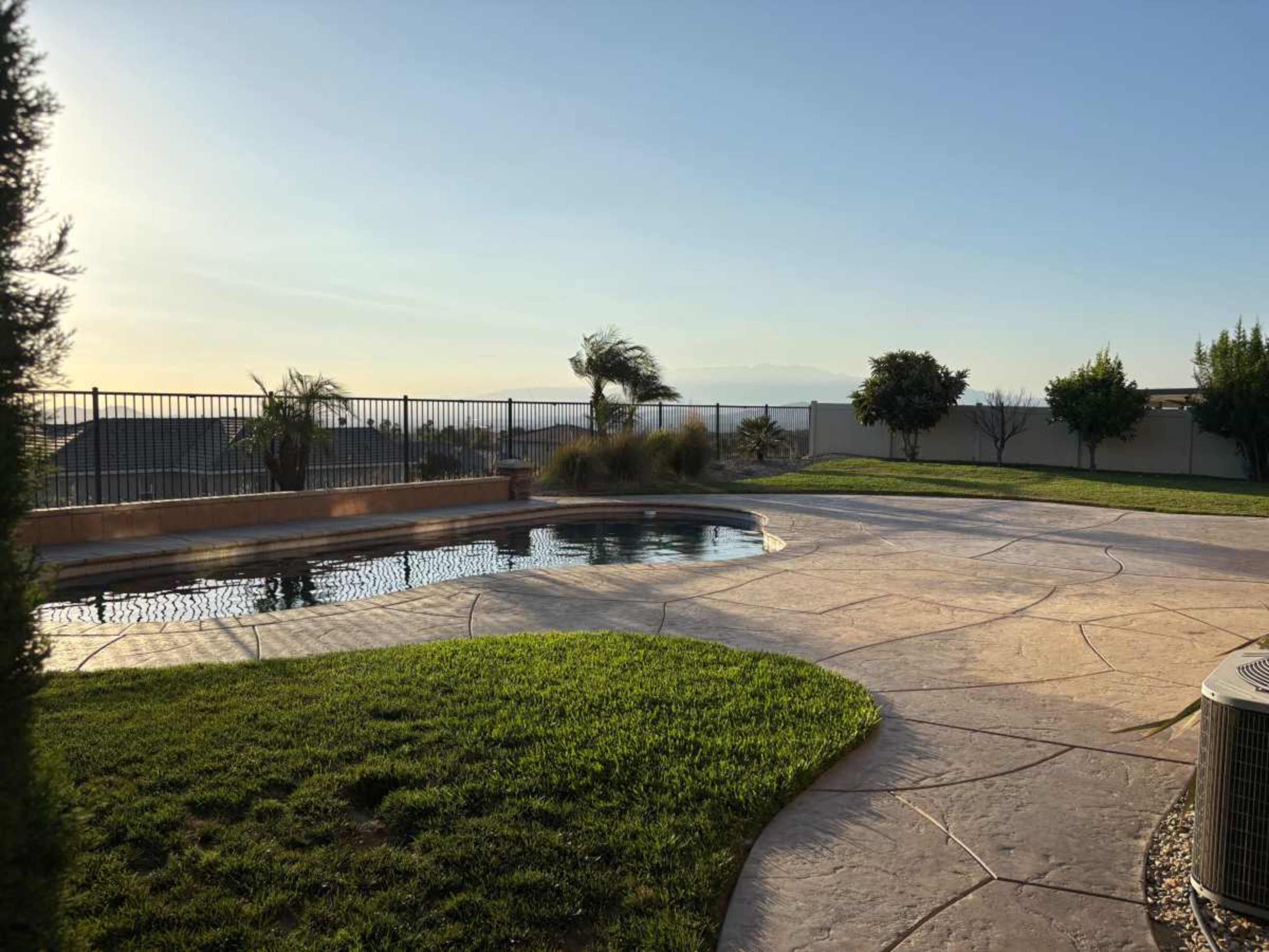 The image shows a landscaped backyard with a swimming pool, surrounded by grass and a fence, under a clear sky.