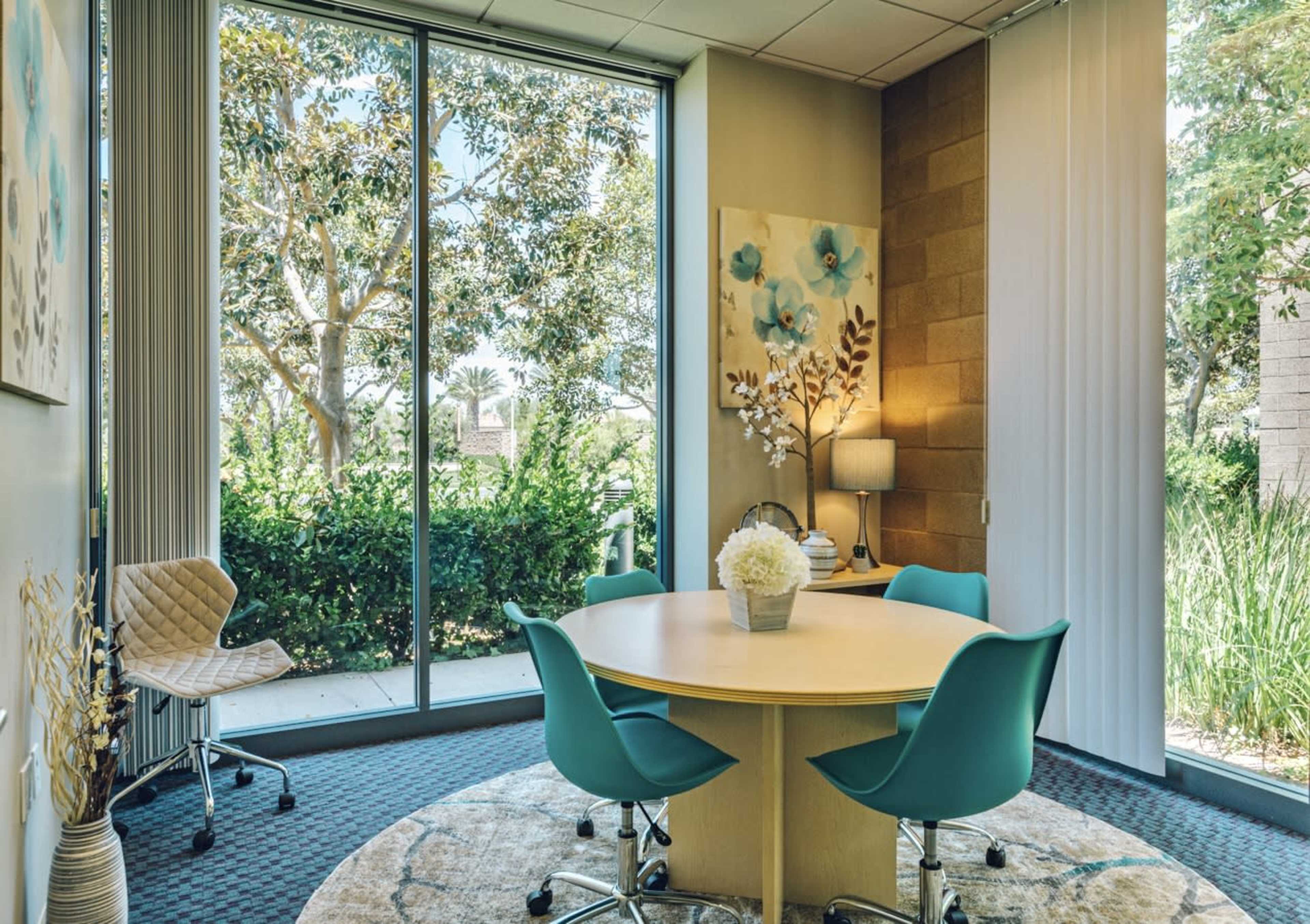 A small meeting room features a round wooden table surrounded by teal chairs, with large windows offering a view of greenery outside.