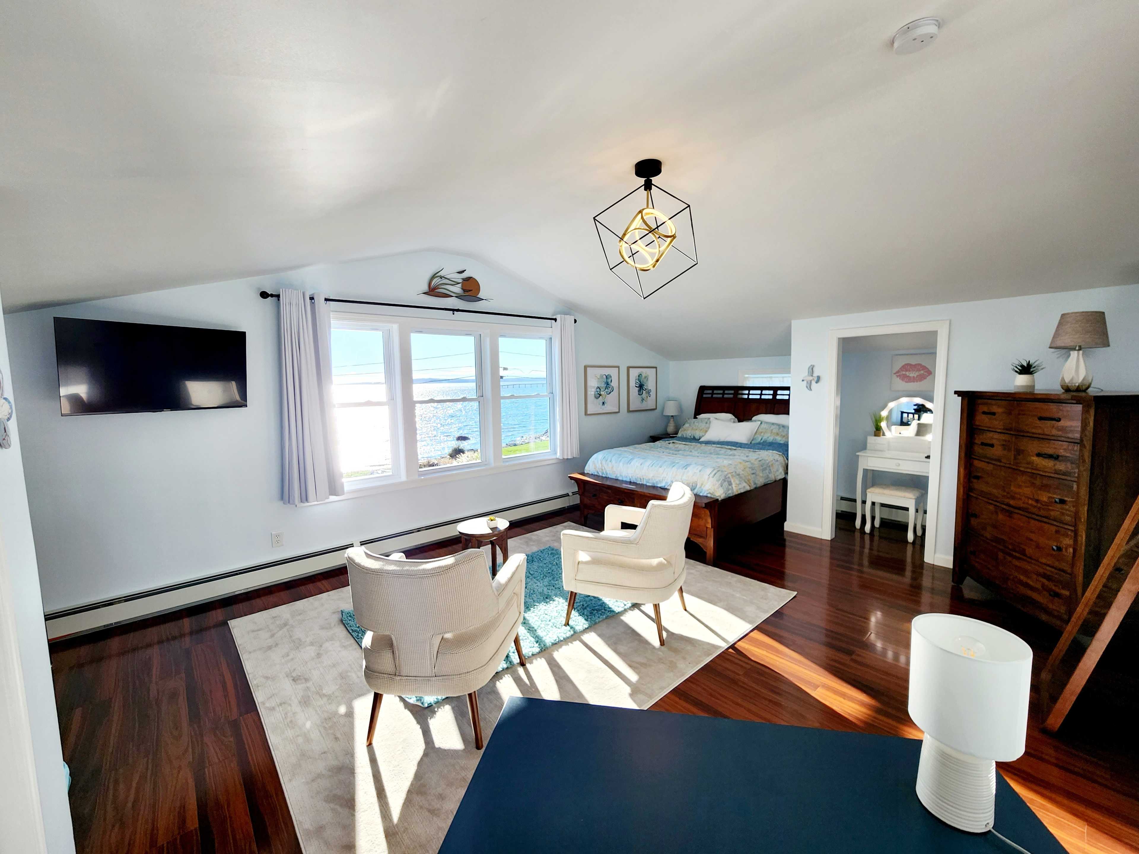 A bright, airy bedroom with large windows offering a view of the ocean, featuring a bed, two chairs, a dresser, and a television.