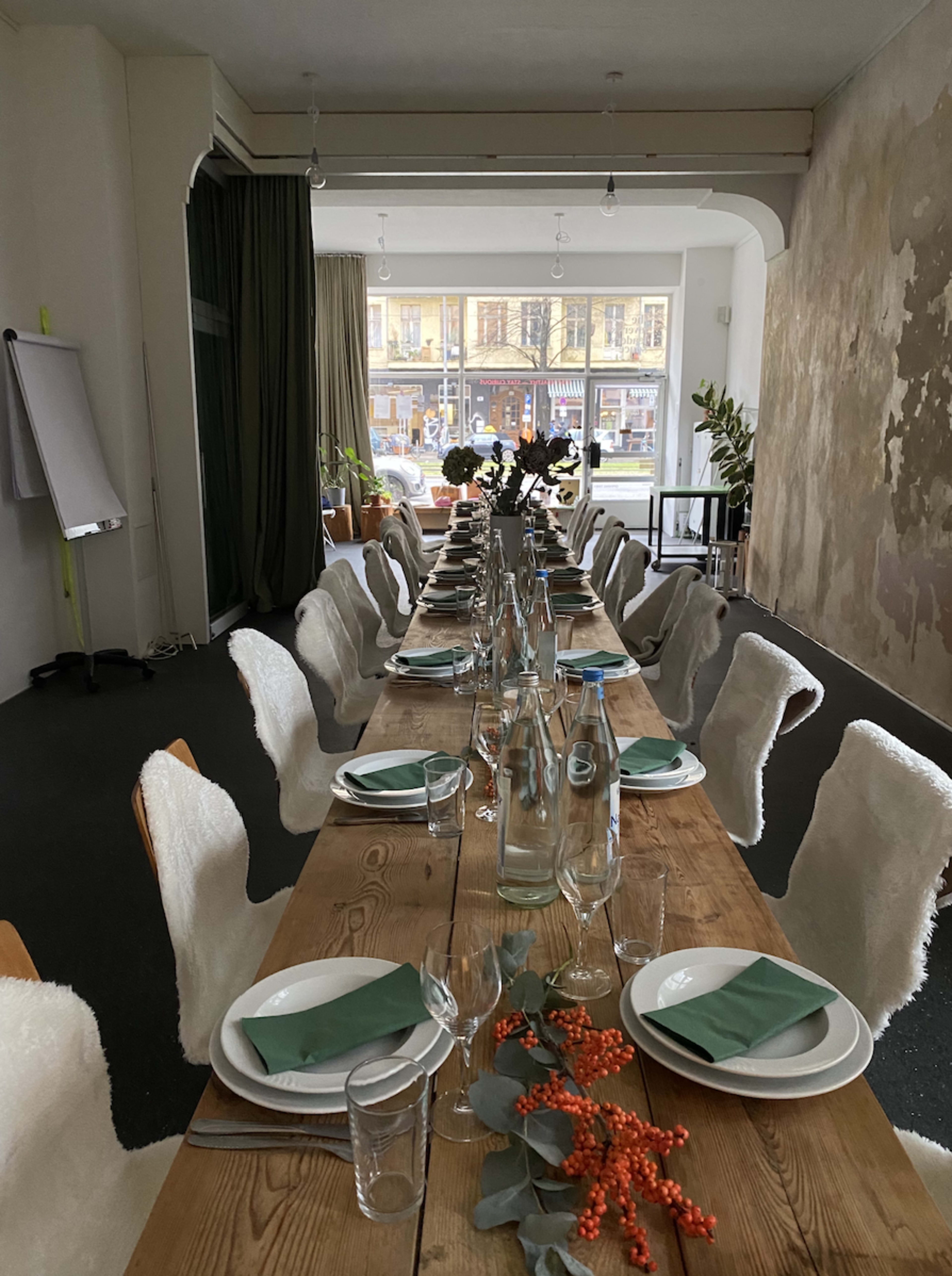 A long wooden dining table is set with plates, glasses, and decorative greenery in a spacious room with large windows.
