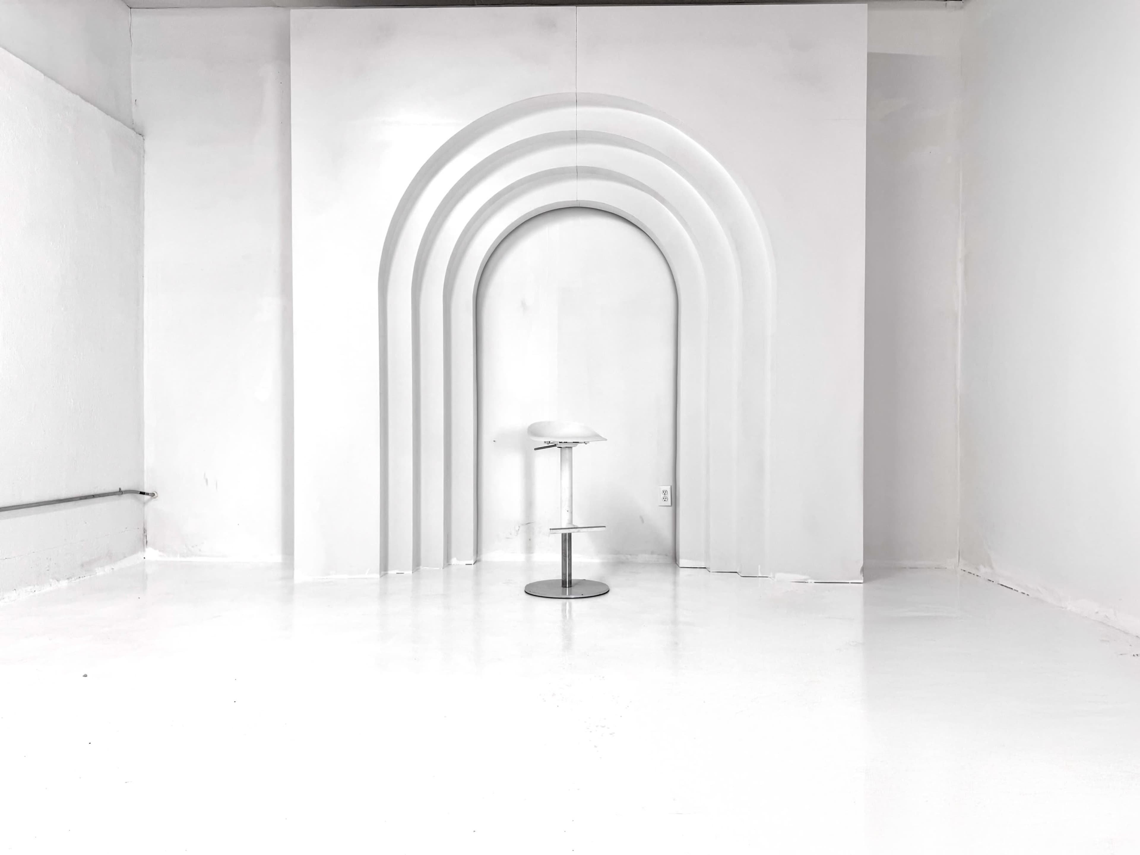 A minimalistic interior features a white wall with curved arch designs and a solo stool in the center.
