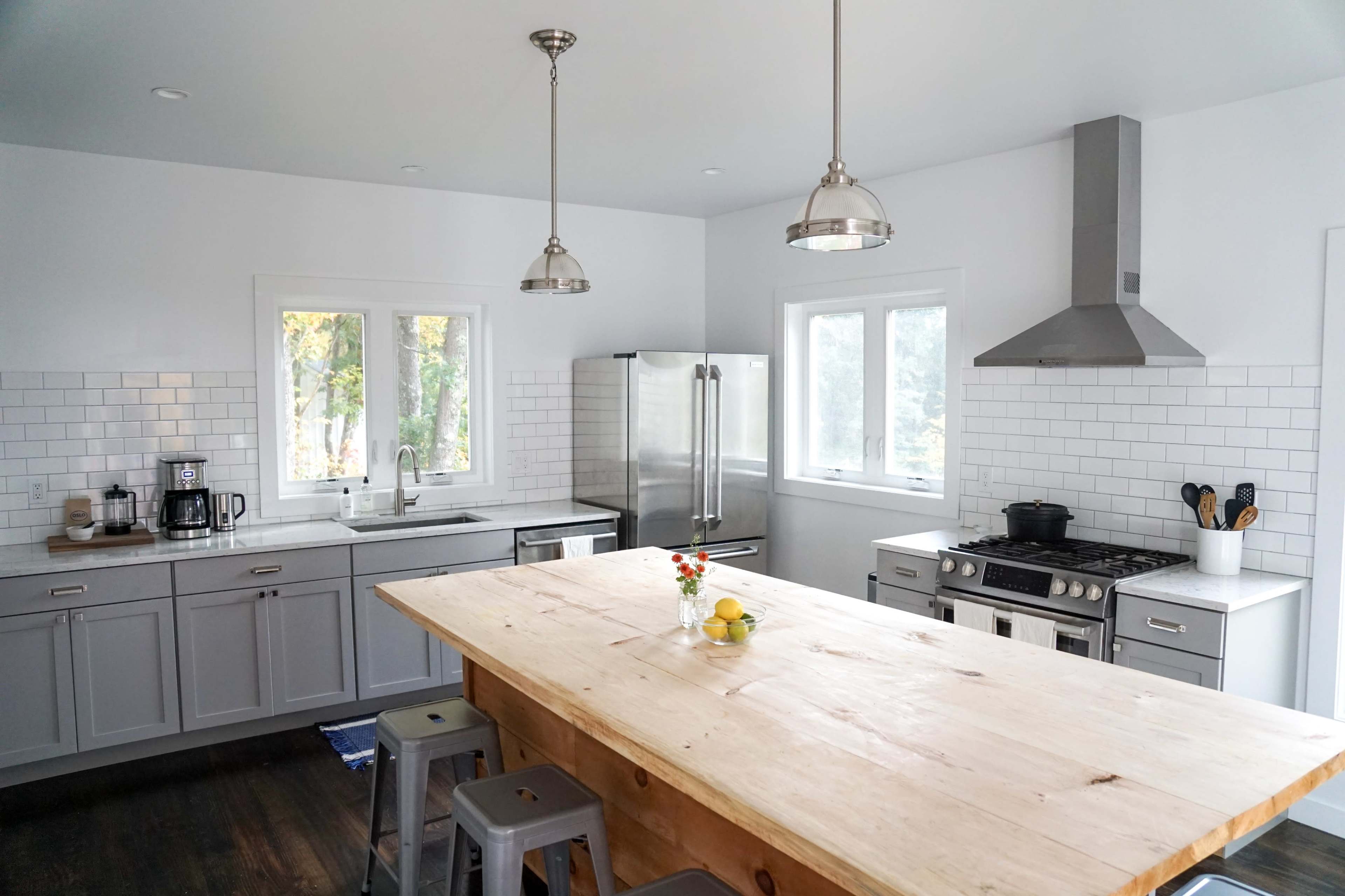 A modern kitchen features a large wooden island, stainless steel appliances, gray cabinets, and white tile backsplash.