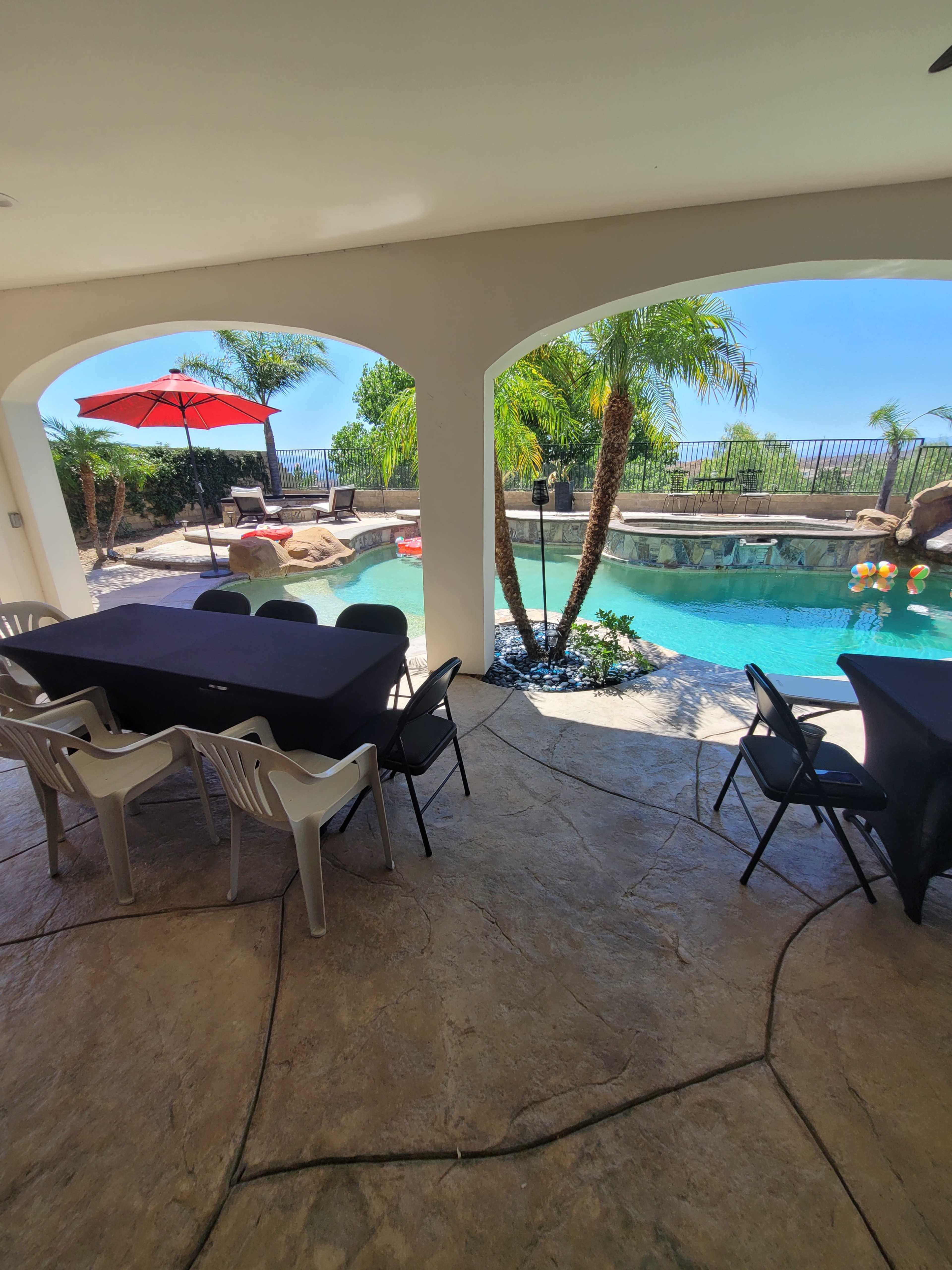 A shaded outdoor area with a large table and chairs overlooking a swimming pool surrounded by palm trees and a rock formation.