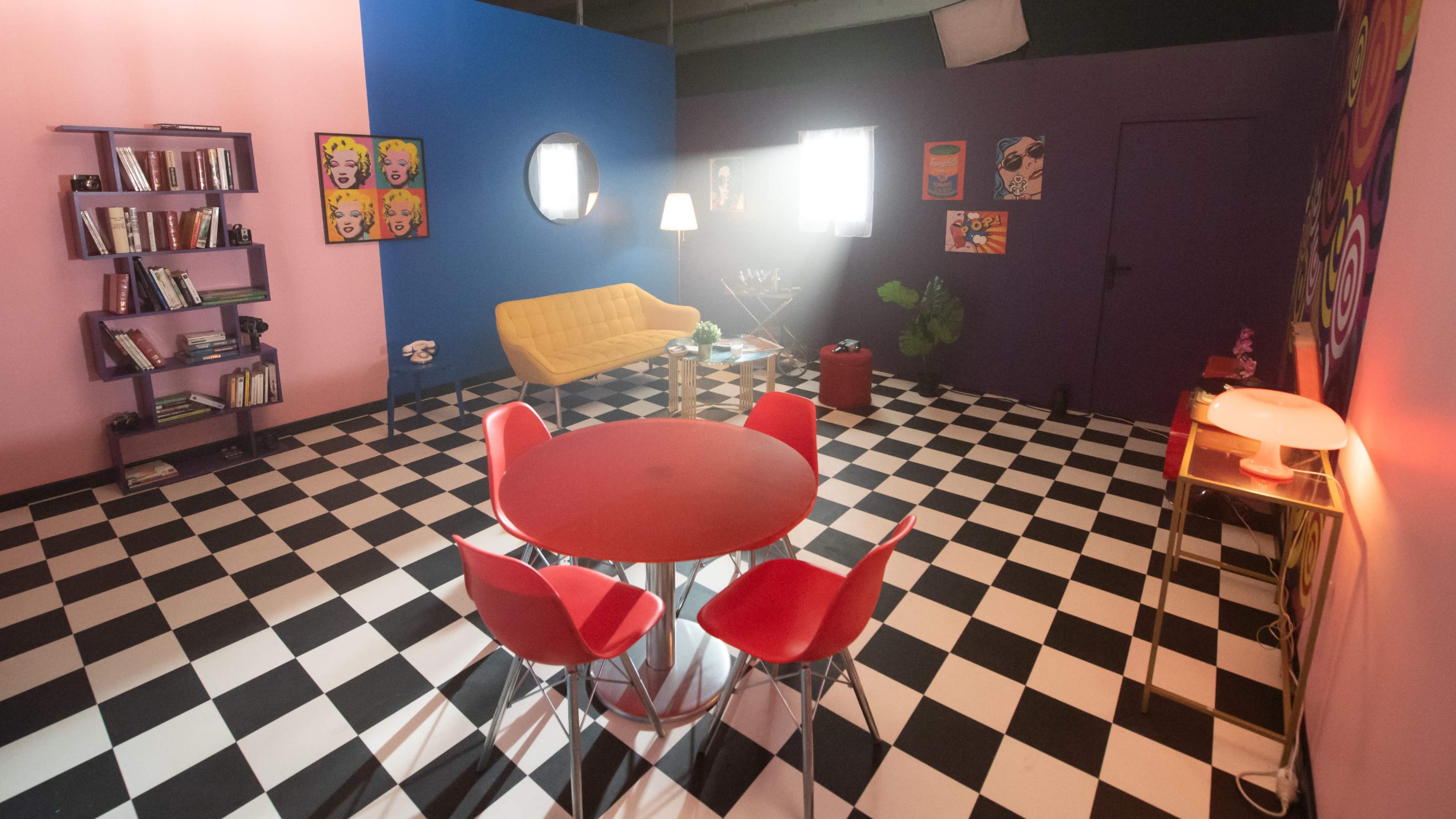 The room features a checkered black and white floor, a yellow sofa, a round red dining table surrounded by red chairs, and colorful wall decorations.