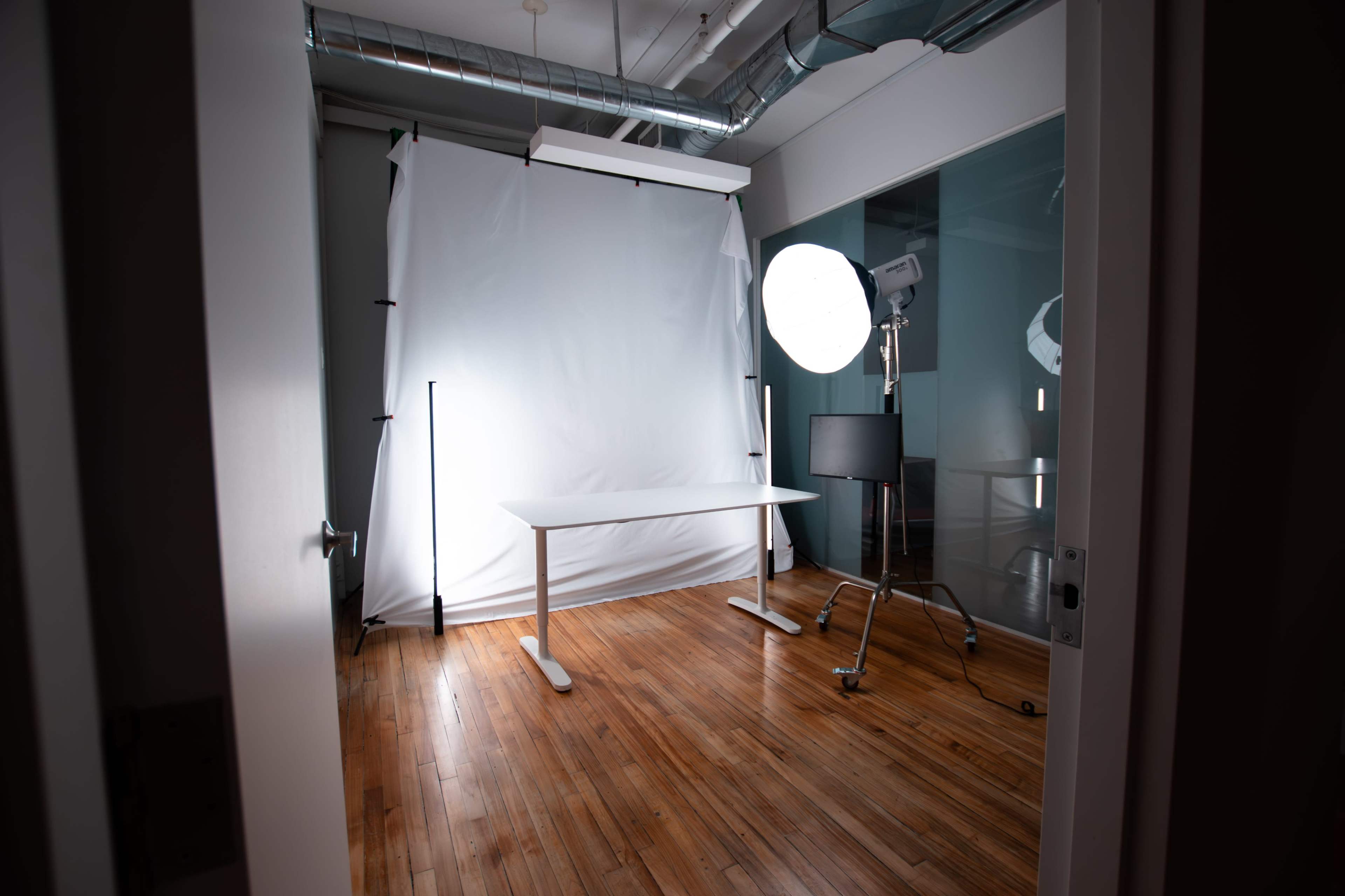The image shows a photography studio featuring a white backdrop, a flat table, and a light setup in a room with wooden flooring.