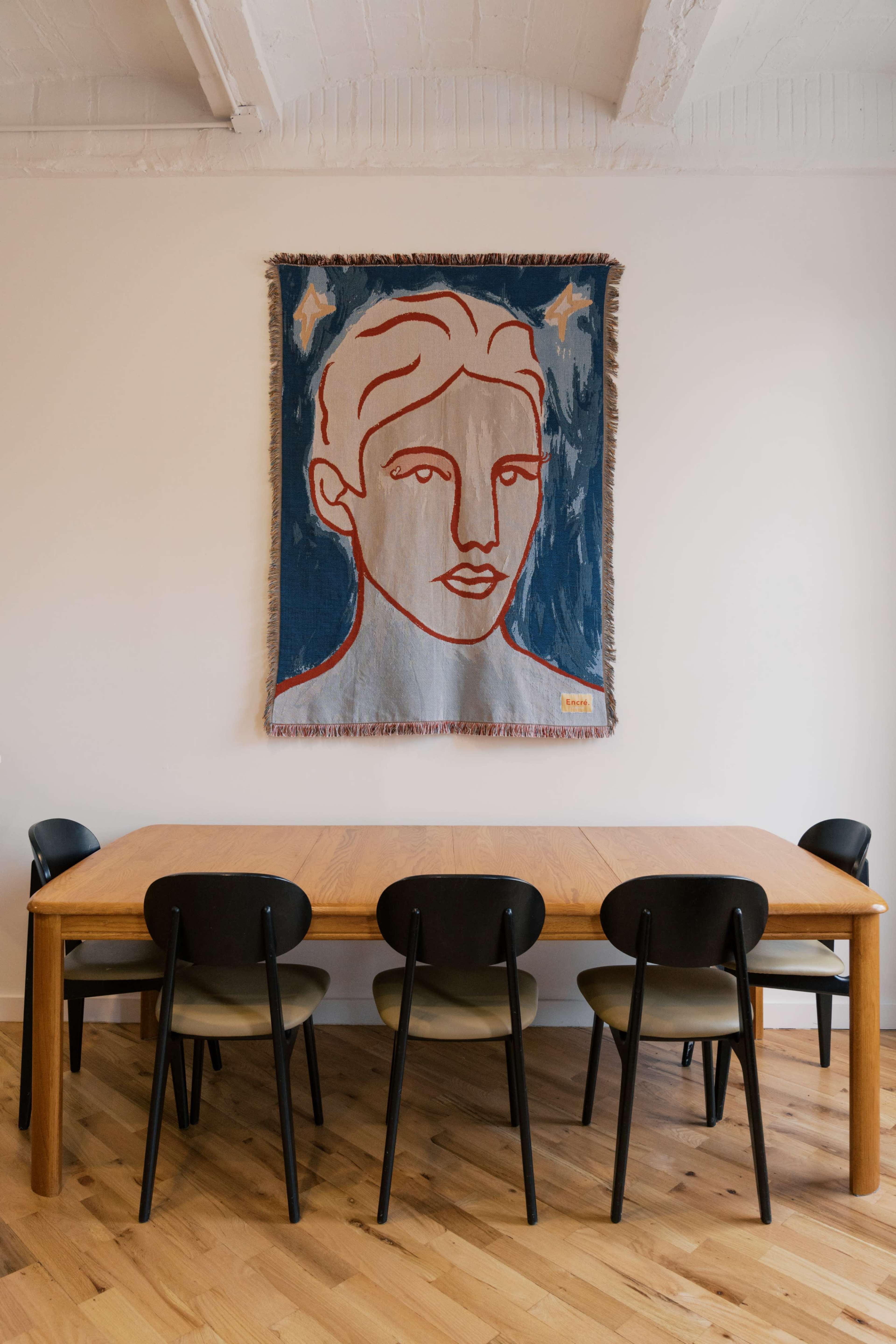 A dining table with six chairs is positioned beneath a woven tapestry featuring a stylized portrait on a blue background.