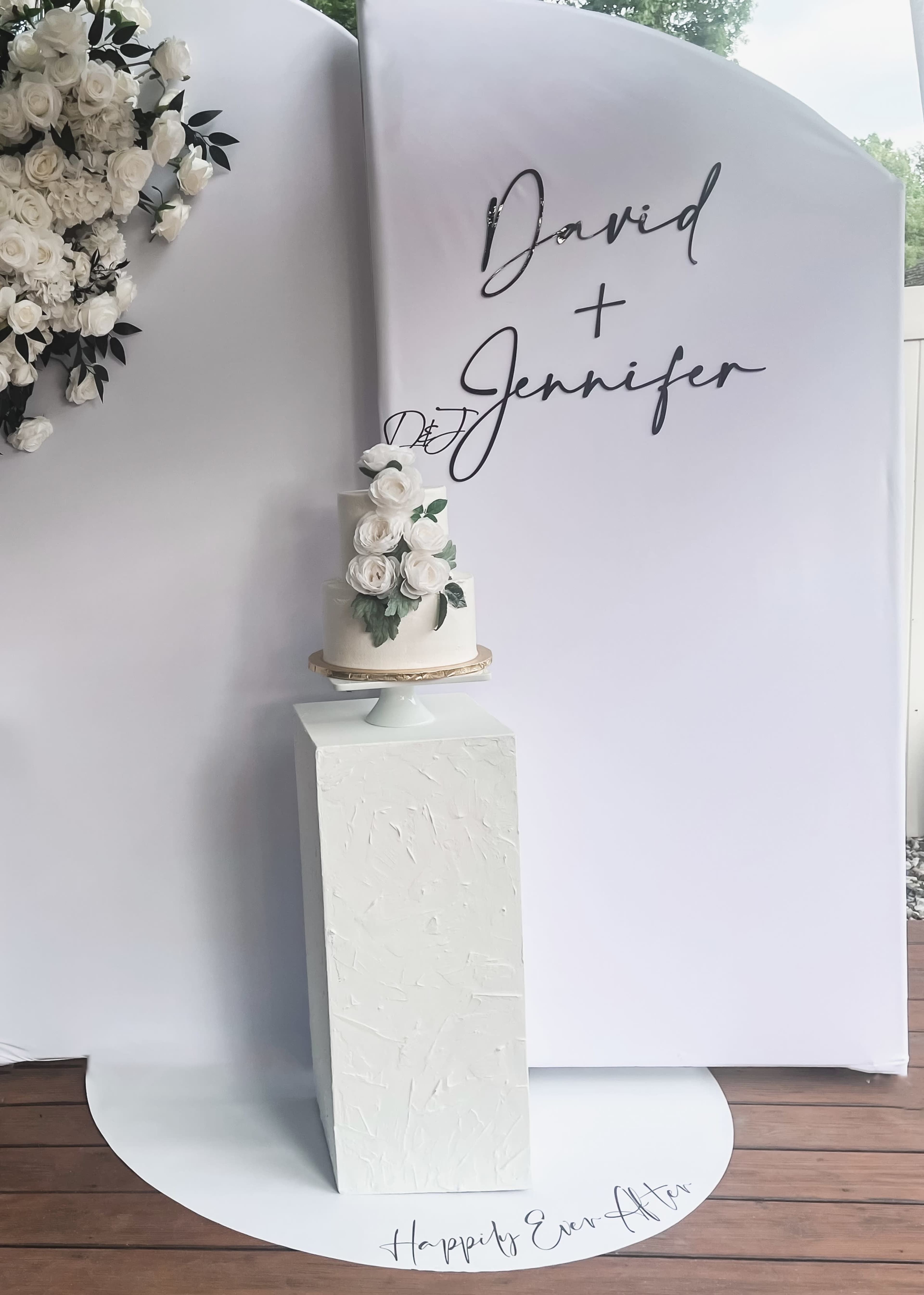 A wedding cake adorned with white flowers is displayed on a pedestal in front of a backdrop featuring the names "David" and "Jennifer."