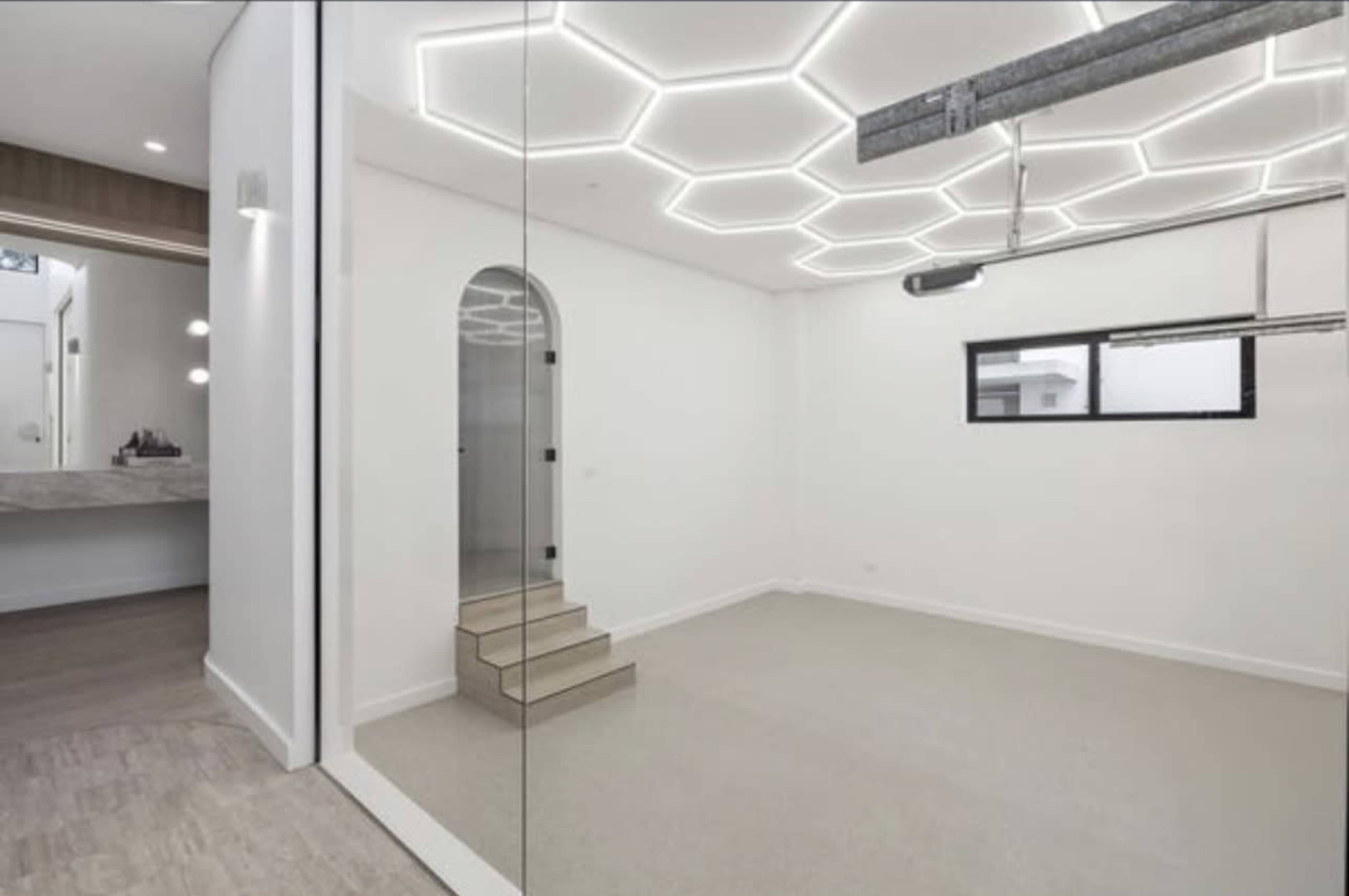The image depicts a modern, minimalistic room with a geometric ceiling design, stairs leading to a different level, and large windows providing natural light.