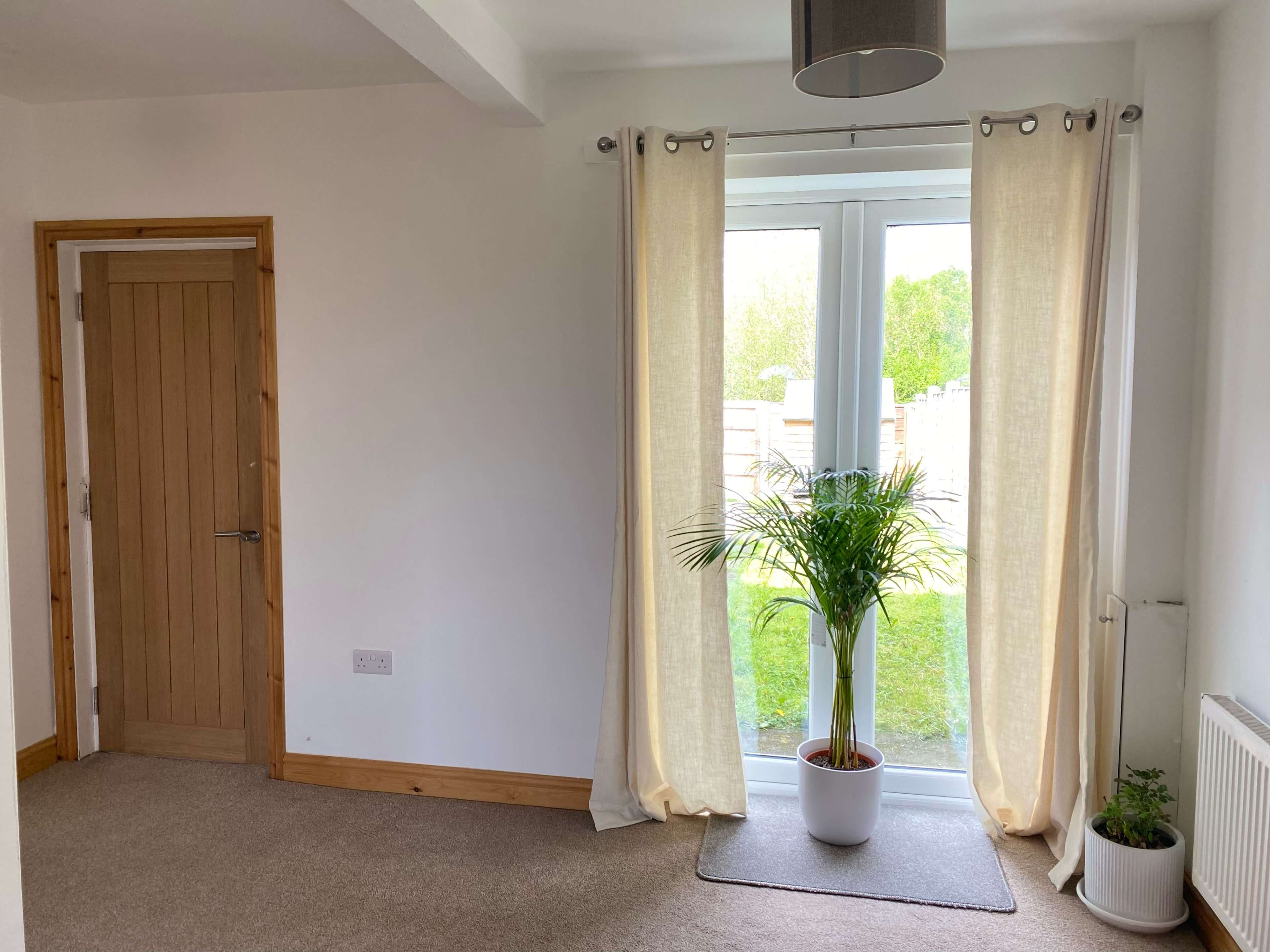 The image shows a corner of a room with a door on the left, a pair of curtains framing a glass door leading to a garden, and a potted plant in front of the door.