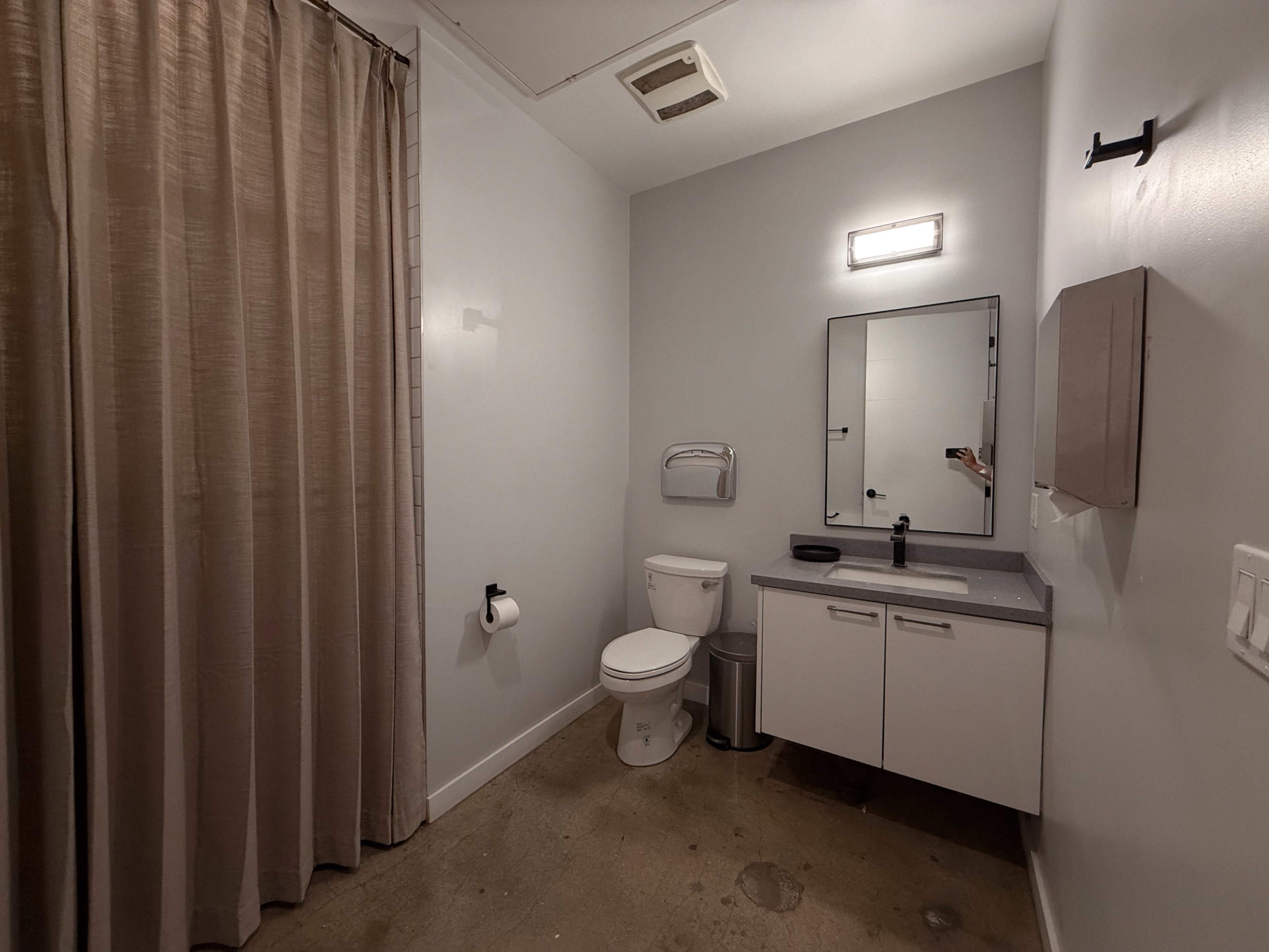 The image shows a minimalist bathroom featuring a toilet, a sink with a mirror above it, a hand towel dispenser, and a shower area separated by a curtain.