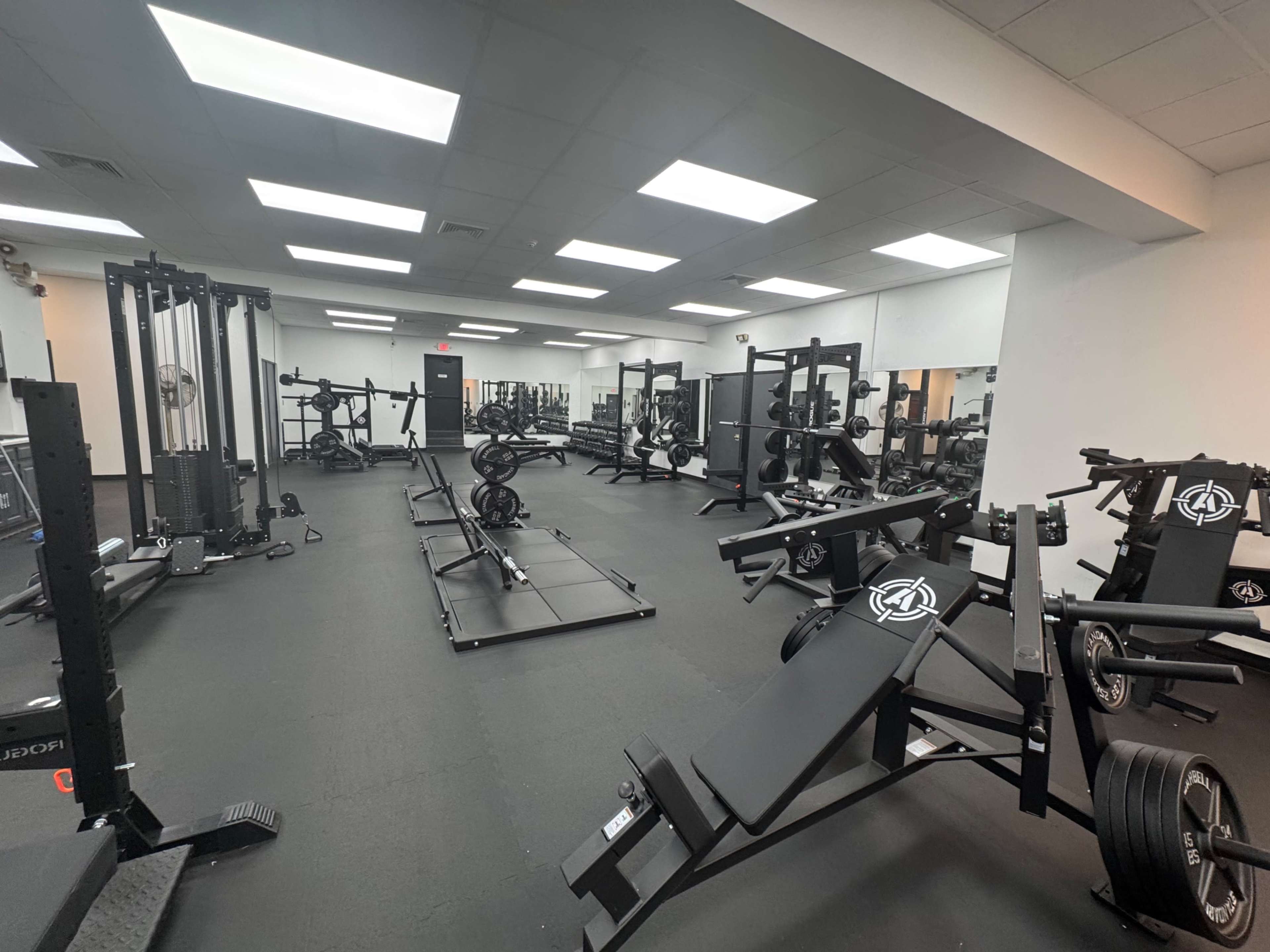 The image shows an empty gym interior filled with various weightlifting equipment and benches arranged on a rubberized floor.