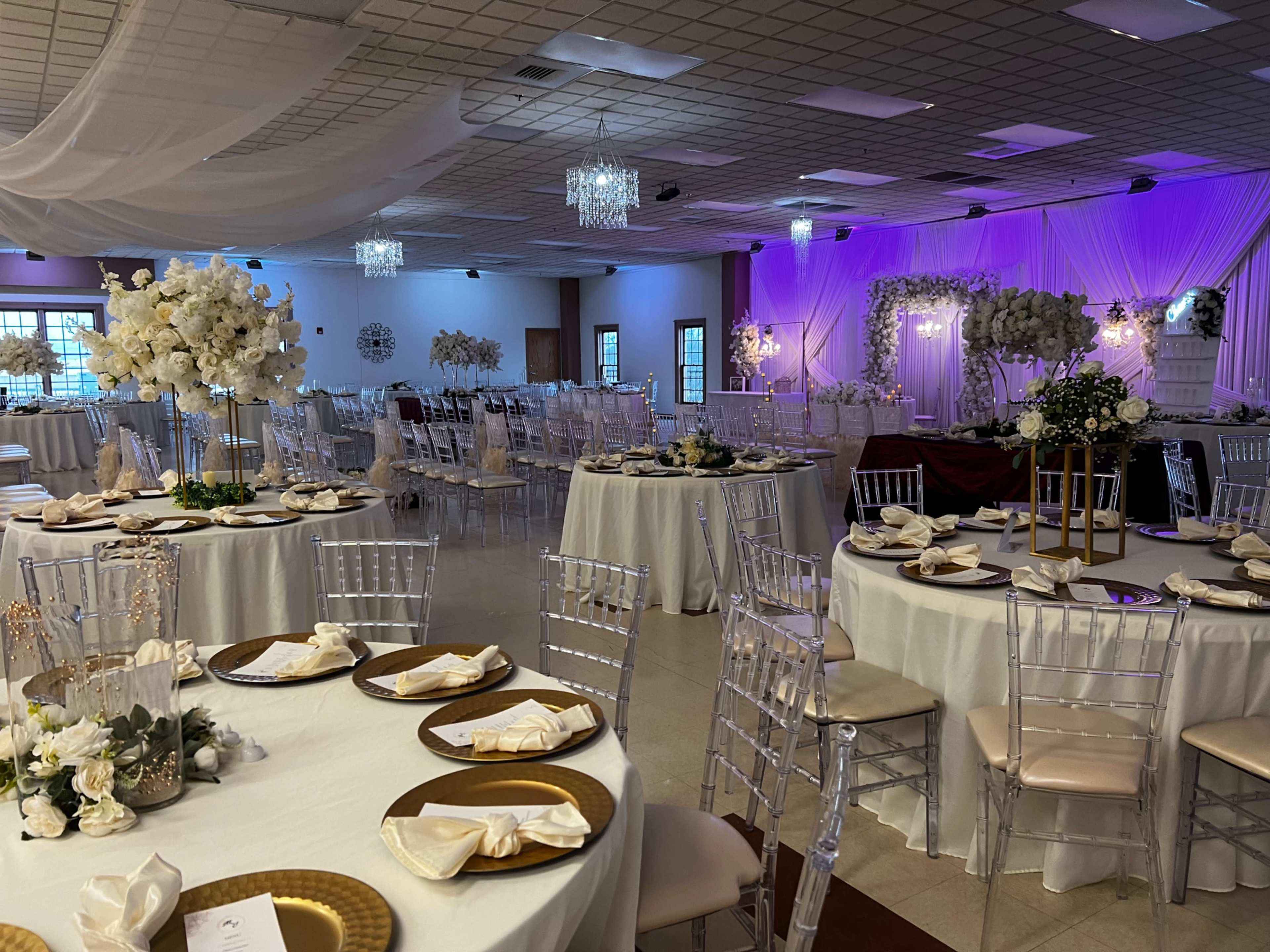 The image shows an elegantly decorated banquet hall with round tables set for an event, adorned with white floral arrangements, gold accents, and ambient lighting.