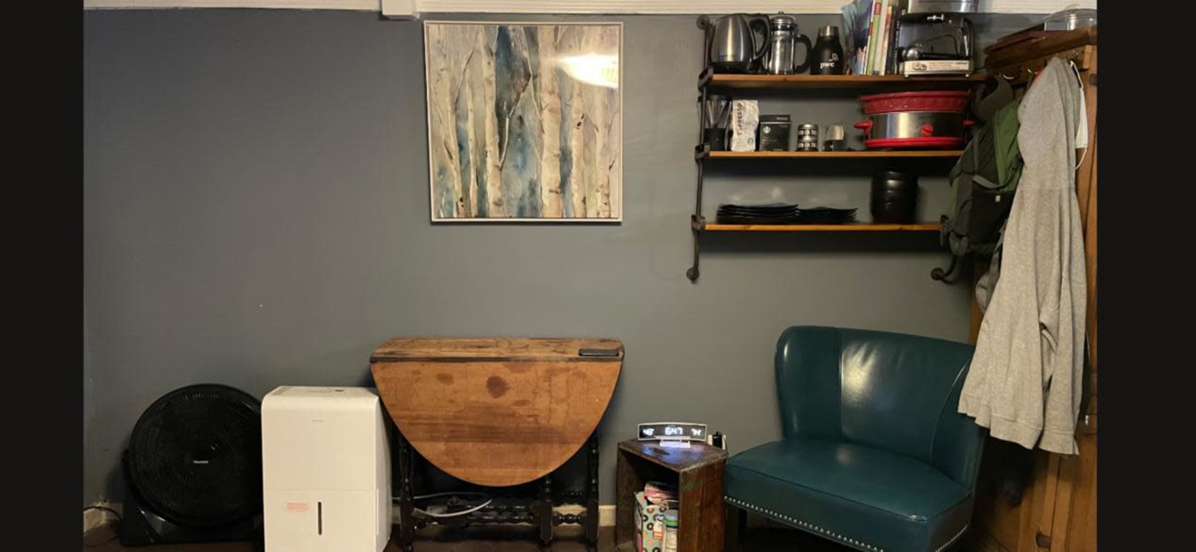 A room features a wall with a painting, a wooden table, a blue chair, a fan, an air conditioner, various kitchen items on a shelf, and a clock on a side table.
