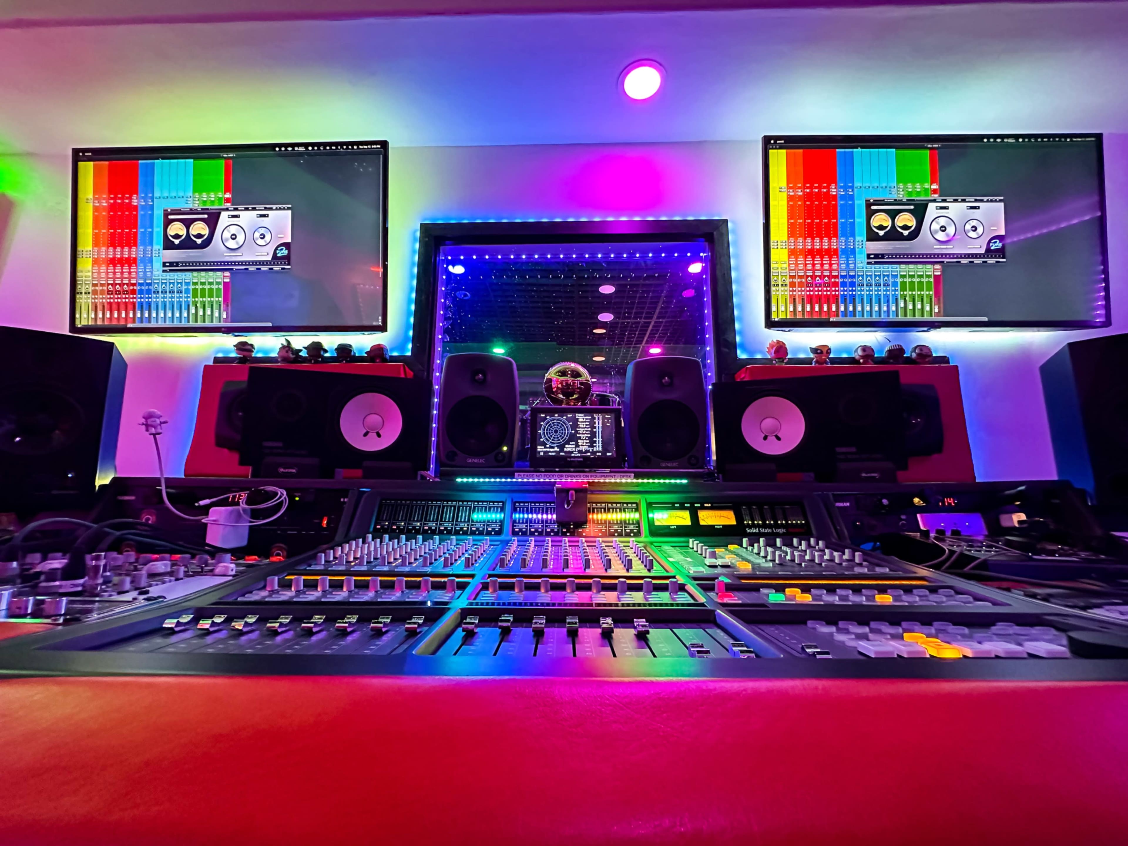 The image shows a music studio with a control desk filled with knobs and sliders, flanked by two large monitors displaying colorful sound wave patterns.