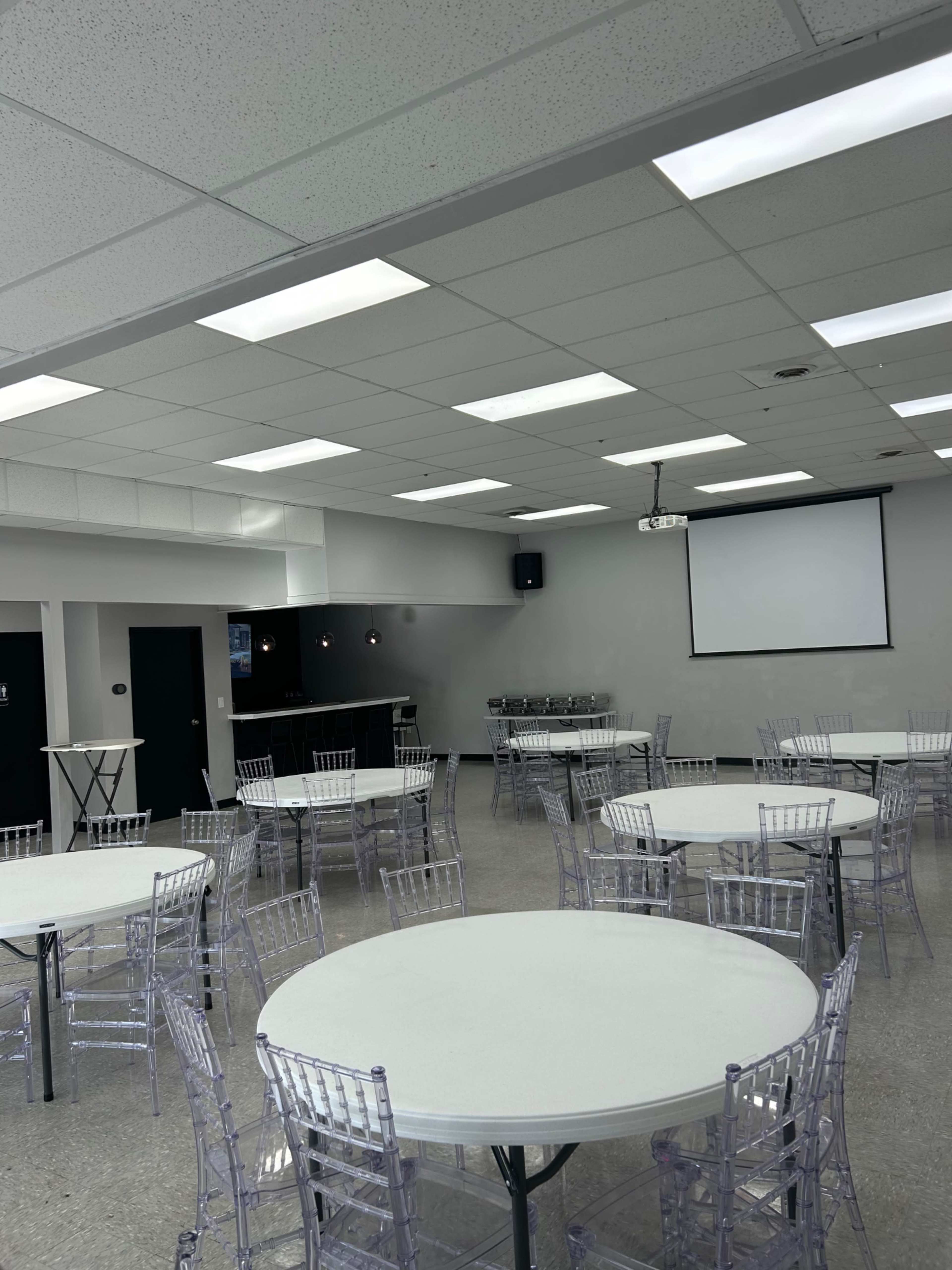 A large event space features several round tables surrounded by clear chairs, with a projector screen and sound equipment mounted on the walls.