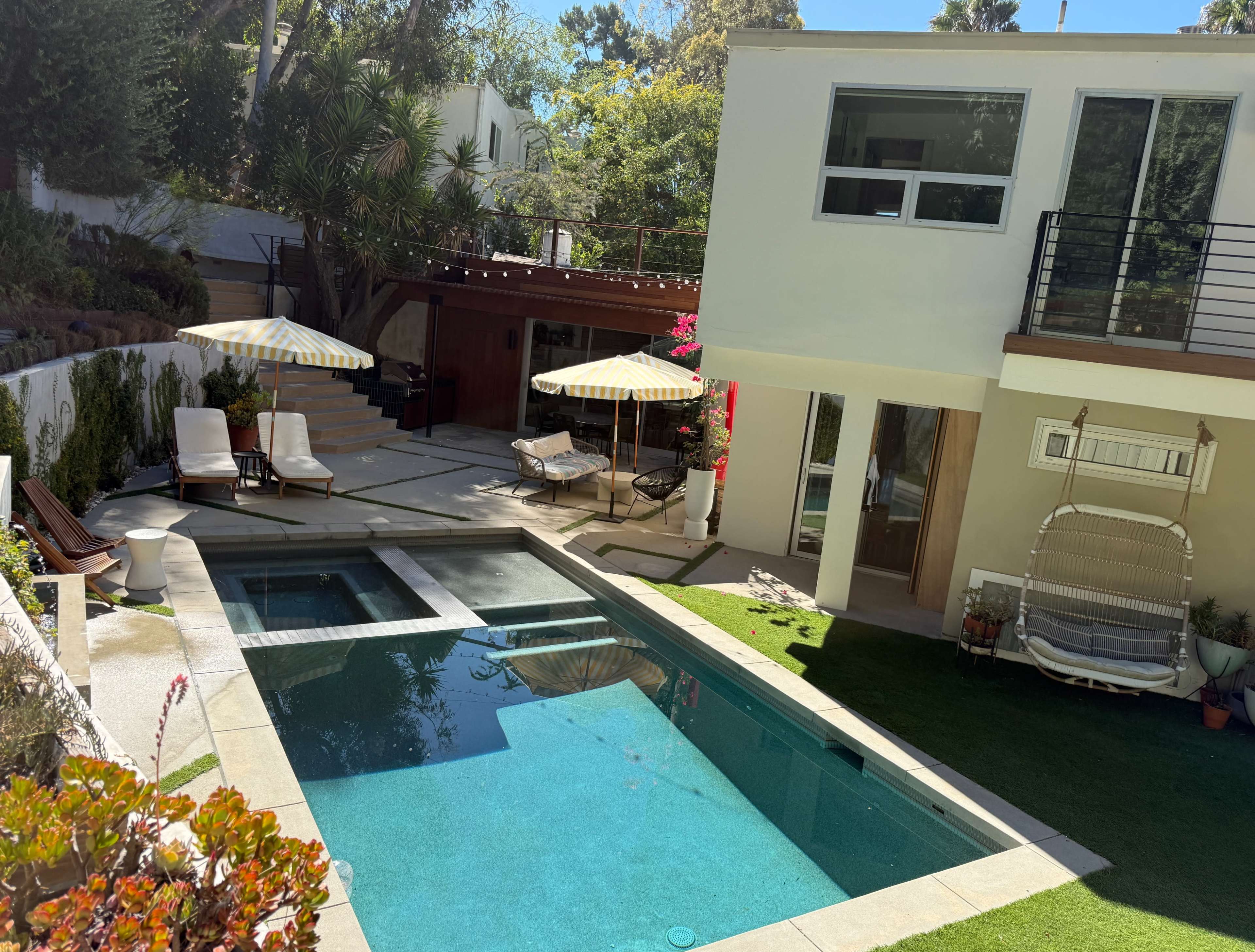 The image shows a modern home with a swimming pool, lounge chairs, and patio umbrellas, surrounded by landscaped gardens.
