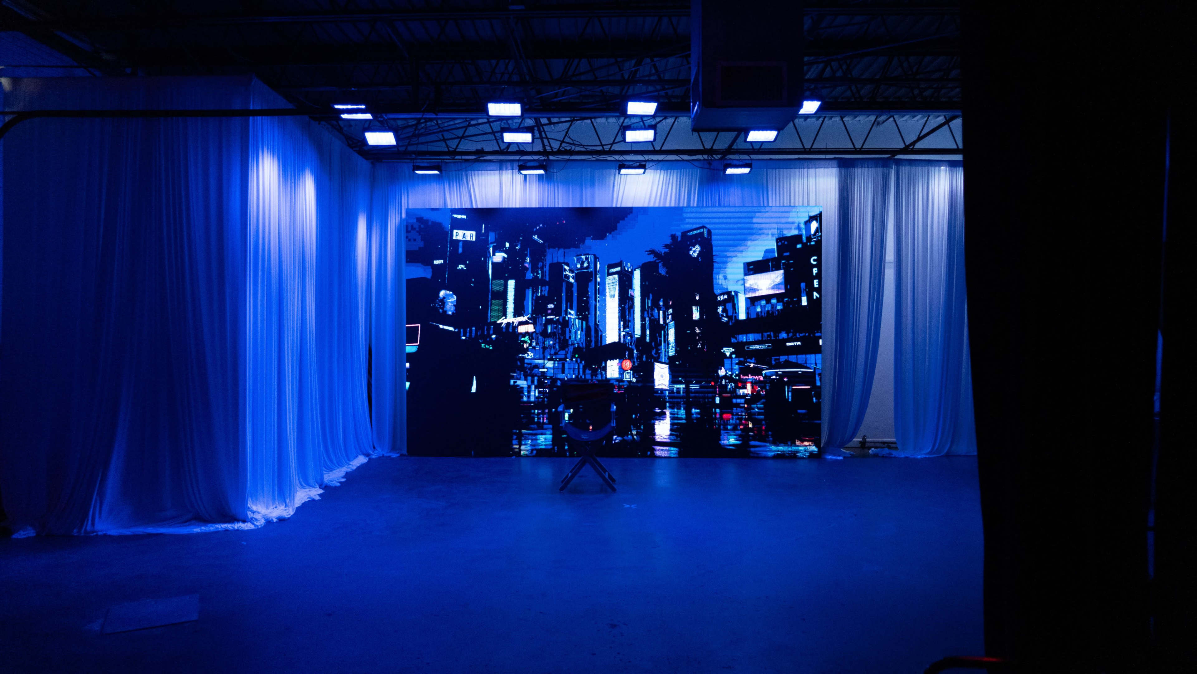 The image shows a studio space with a large, illuminated backdrop depicting a city skyline at night, surrounded by white draped fabric and blue lighting.