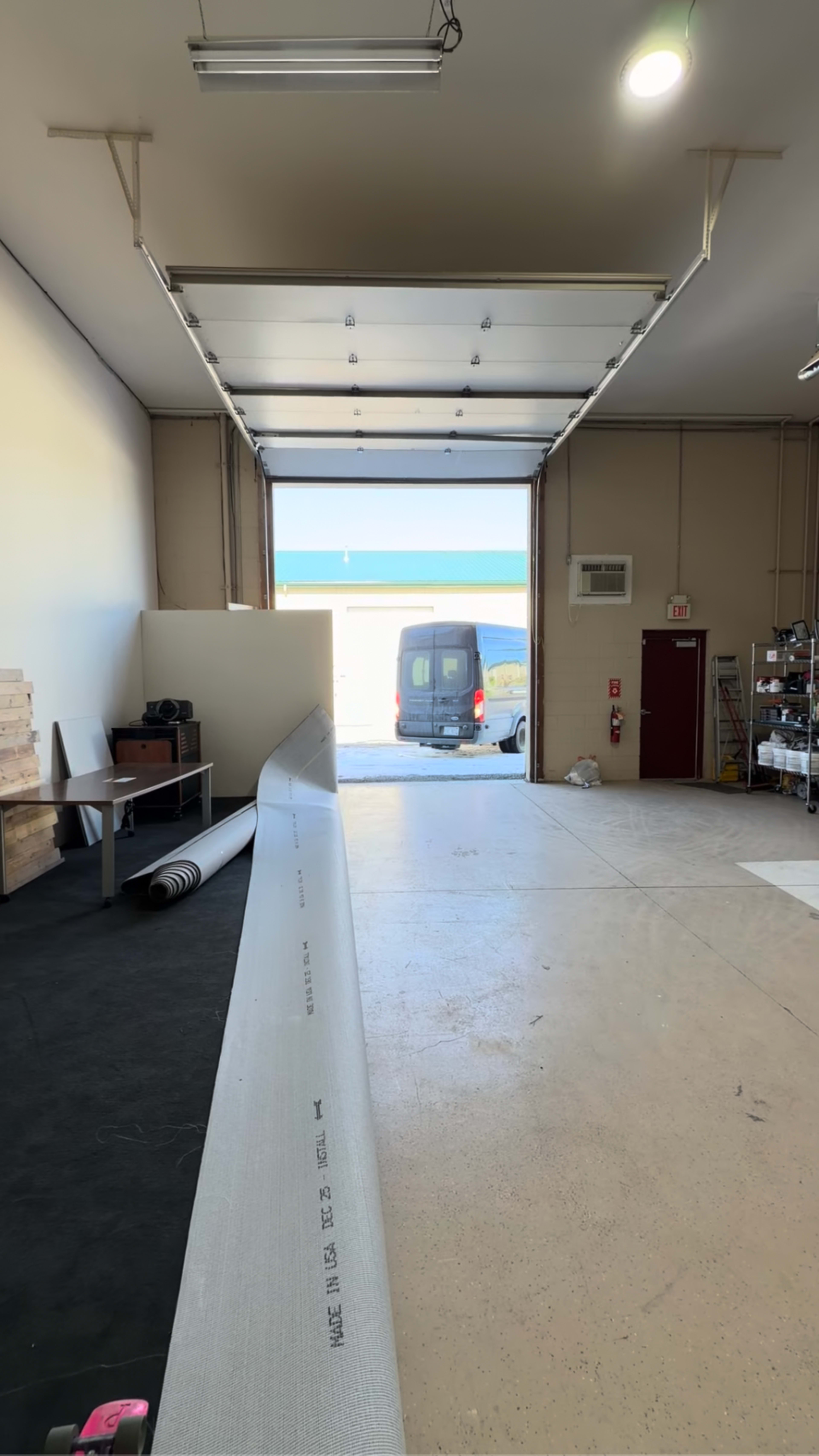 A long piece of material extends along the floor of a spacious garage, with a van visible in the open doorway and daylight streaming in from outside.